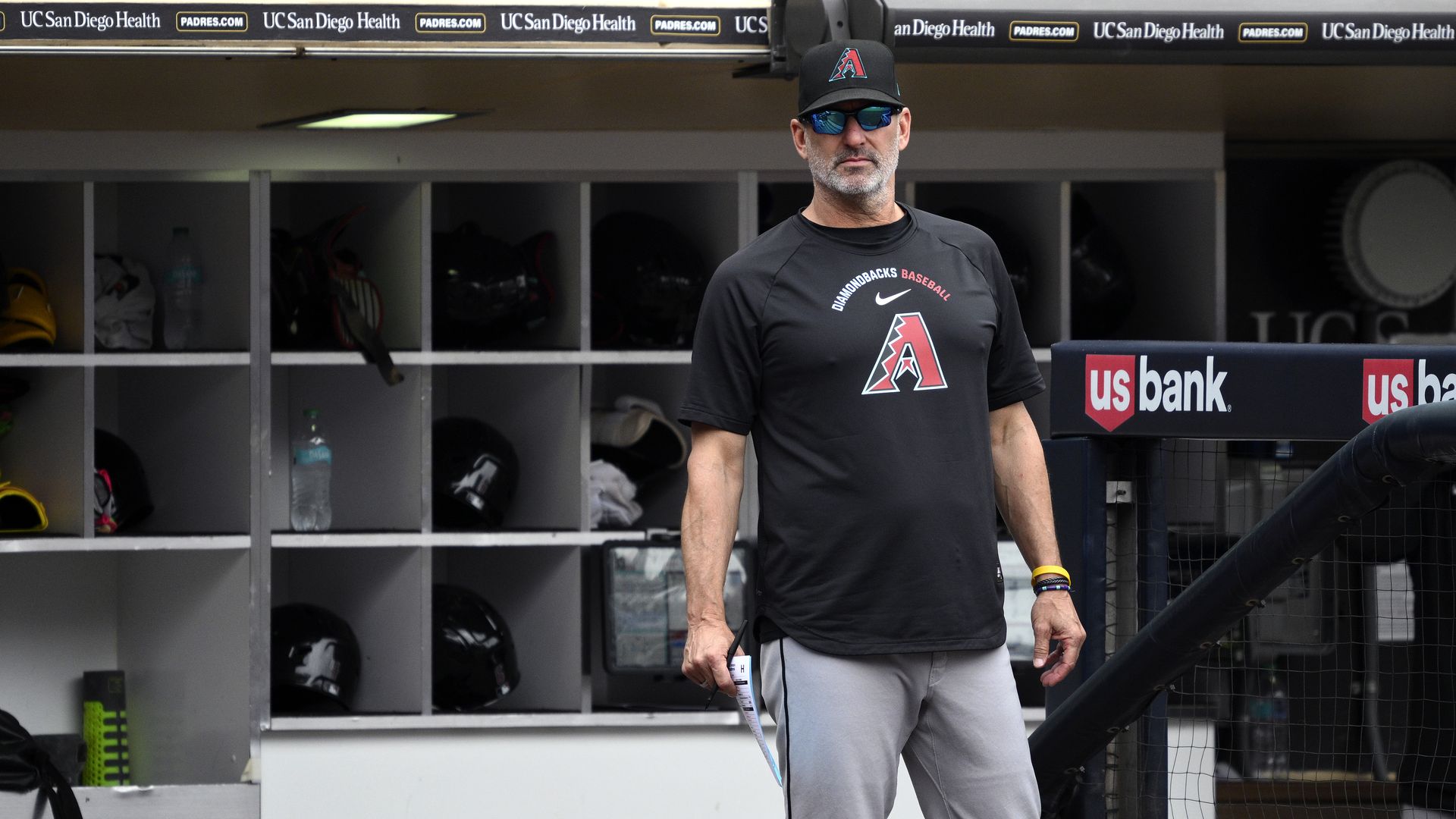 A man in sunglasses, gray pants and a black t-shirt and hat, both with a red "A" on the front, stands in a baseball dugout looking out; cubbyholes filled with various sporting equipment and water bottles are behind him.  