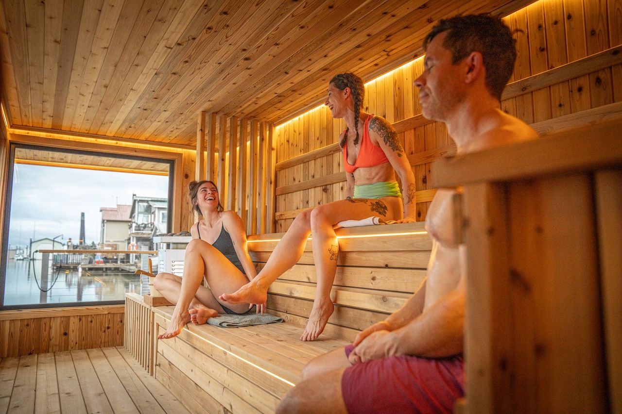 Three people relaxing and smiling inside a wooden sauna with large window view of waterfront buildings and docks in the background.
