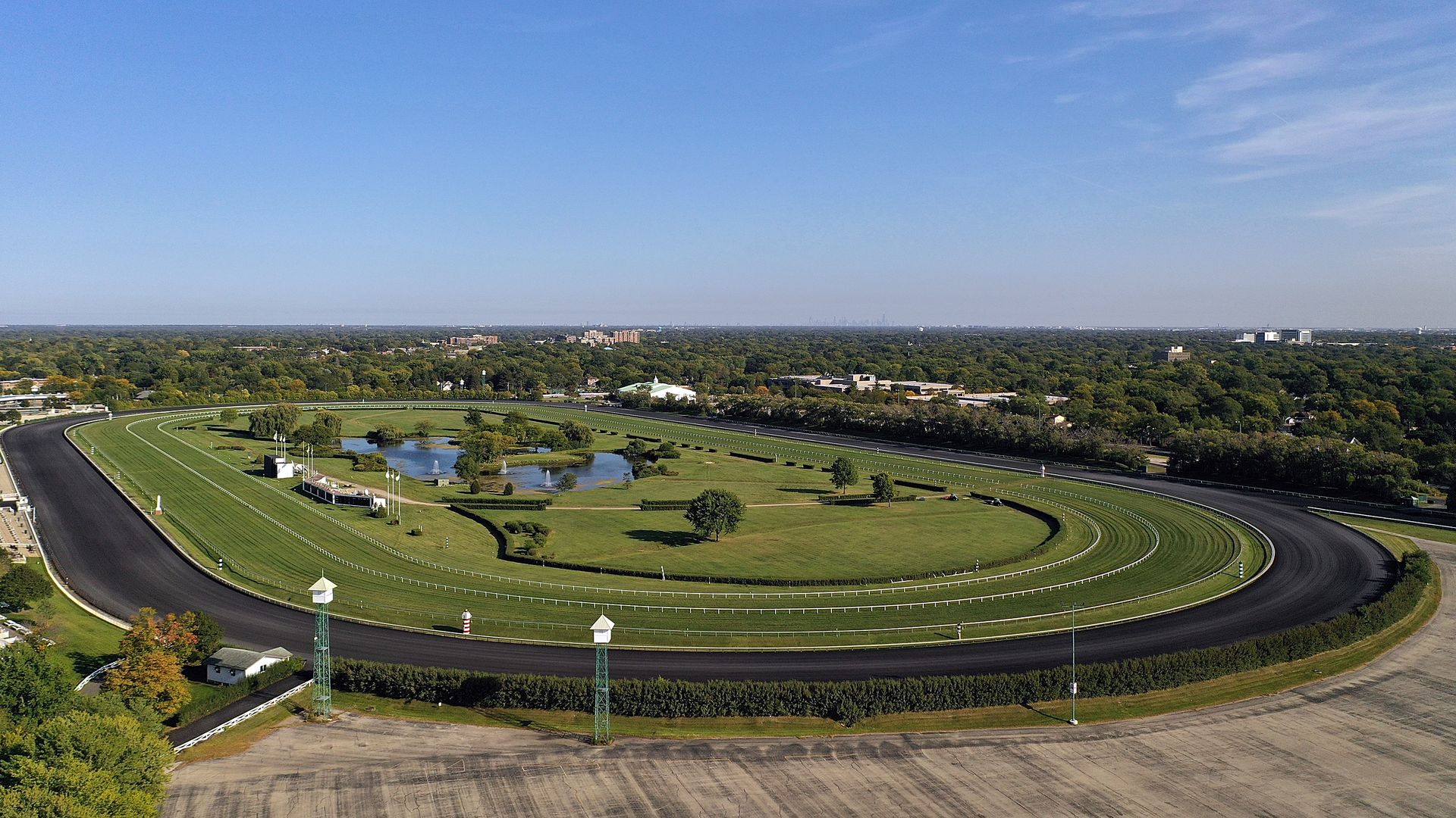 Aerial view of a horse racing track with a dark oval track surrounding manicured green infield and water features. Trees and distant buildings under a clear blue sky.