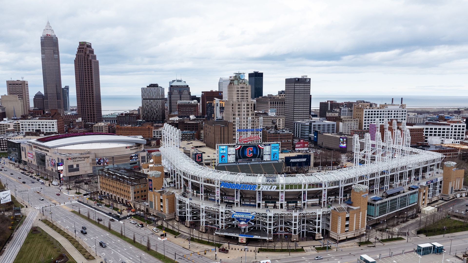 Aerial view of downtown Cleveland with Progressive Field in the foreground, tall office buildings, and Lake Erie beyond under overcast skies.