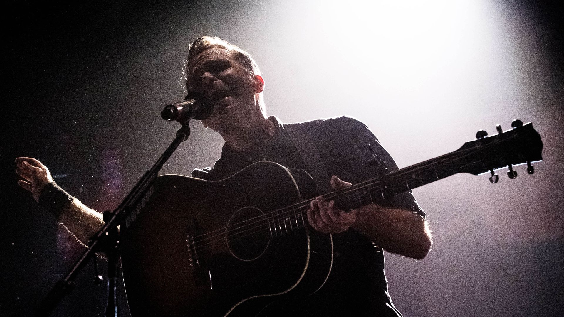 A male singer-guitarist performs on stage, backlit by a bright spotlight to the right. He strums an acoustic guitar and sings into a microphone, wearing a dark shirt.