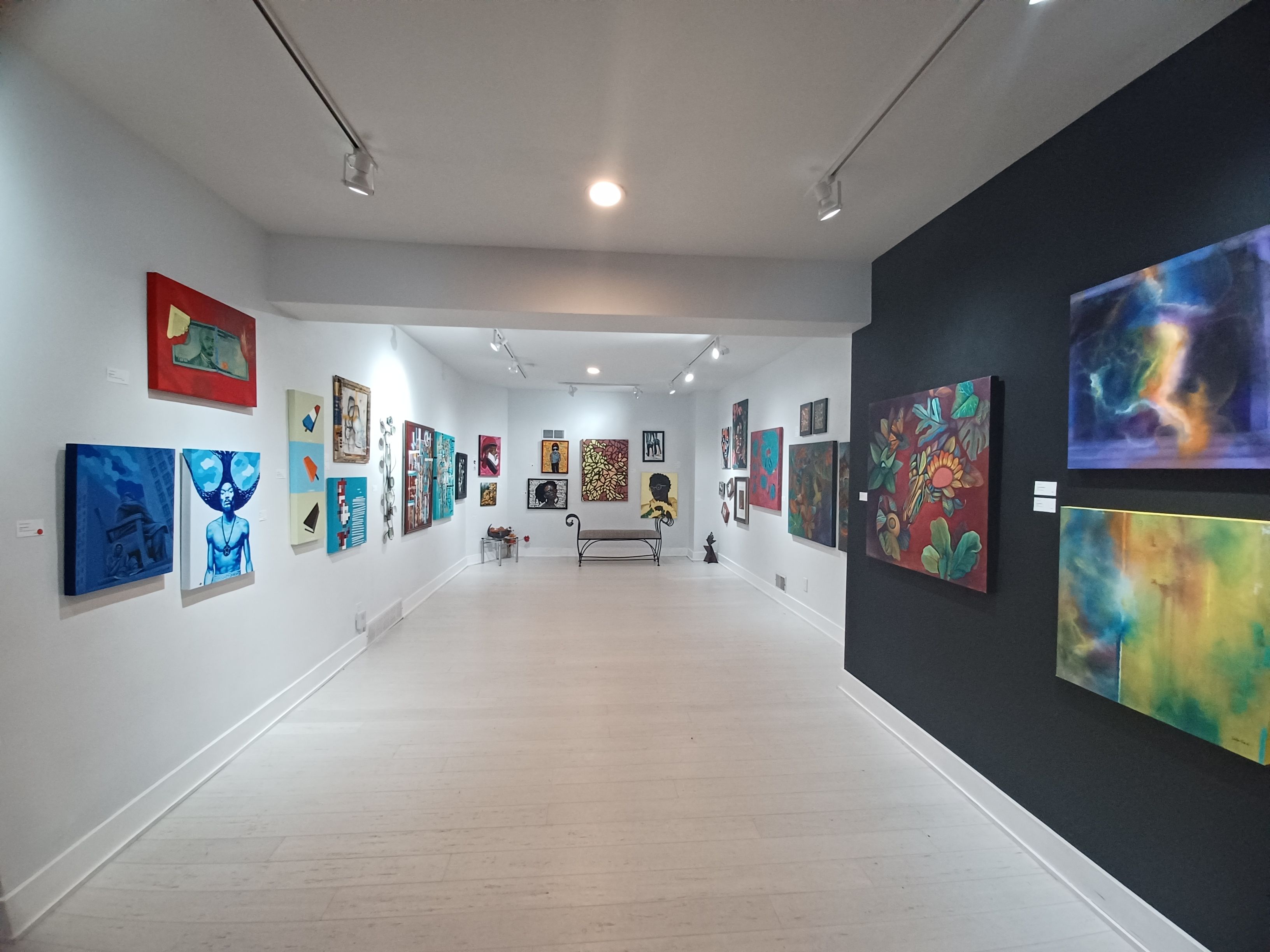 Wide art gallery hallway with white walls and light floor, paintings lining both sides under track lighting, a black accent wall on the right with a vibrant piece, and a bench at the far end.