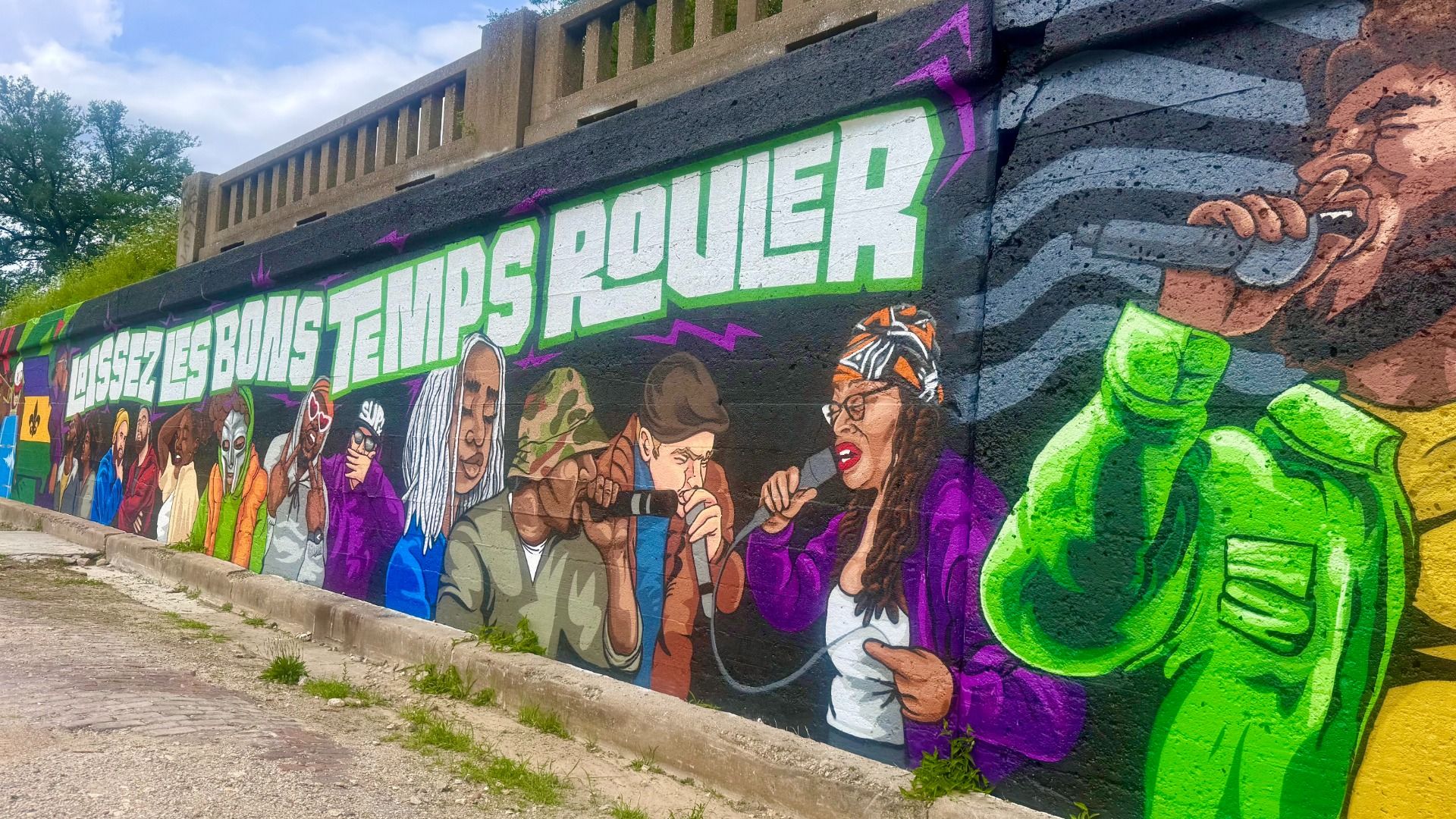 Vibrant mural along a wall features a row of diverse people in colorful clothes, bold white-and-green letters with purple accents, and a bright neon-green figure on the right, under a blue sky.