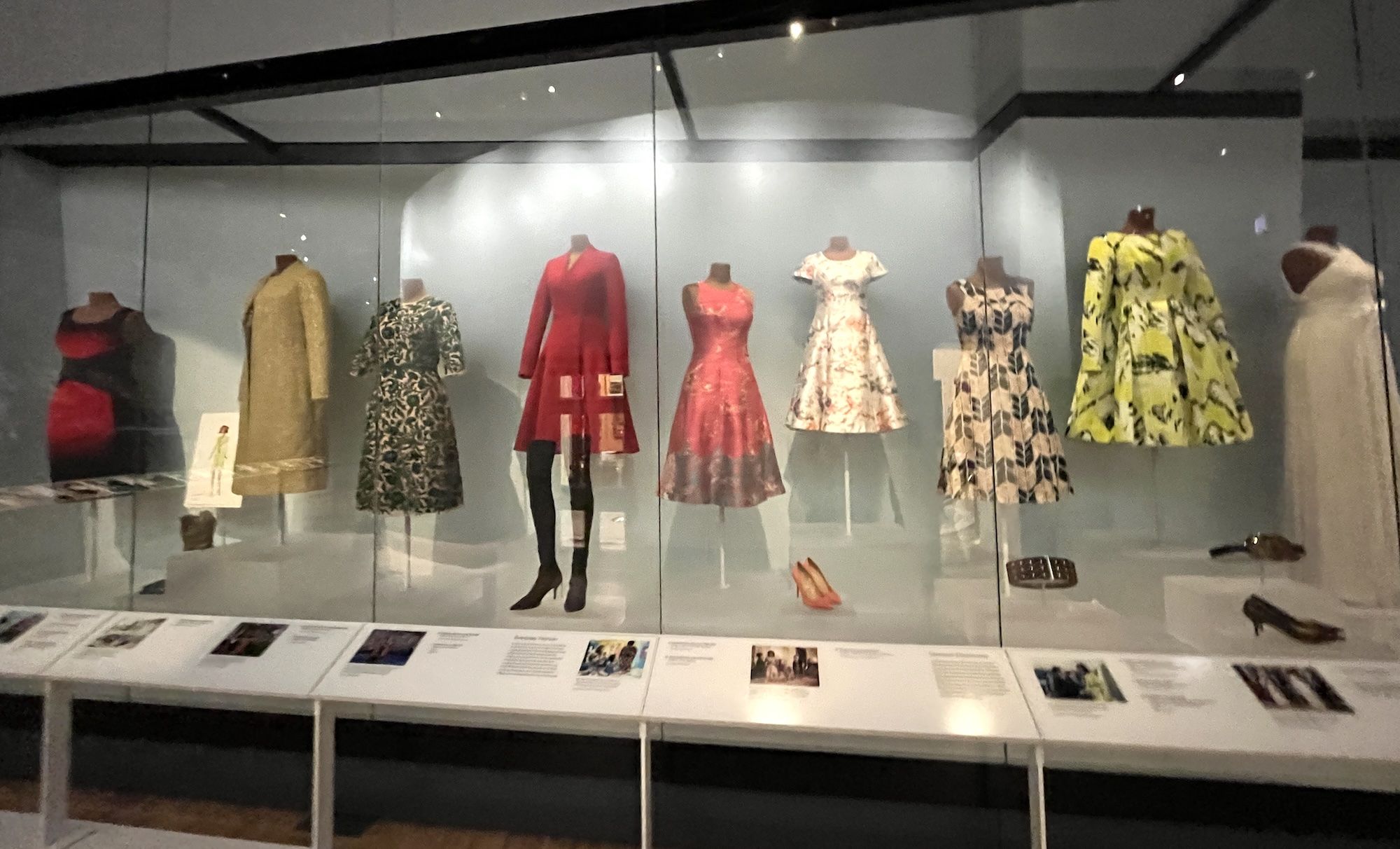 Glass display case with vintage dresses on mannequins: red-black gown, beige coat, green floral dress, red coat, pink dress, white floral dress, blue patterned dress, lime-yellow coat; placards.