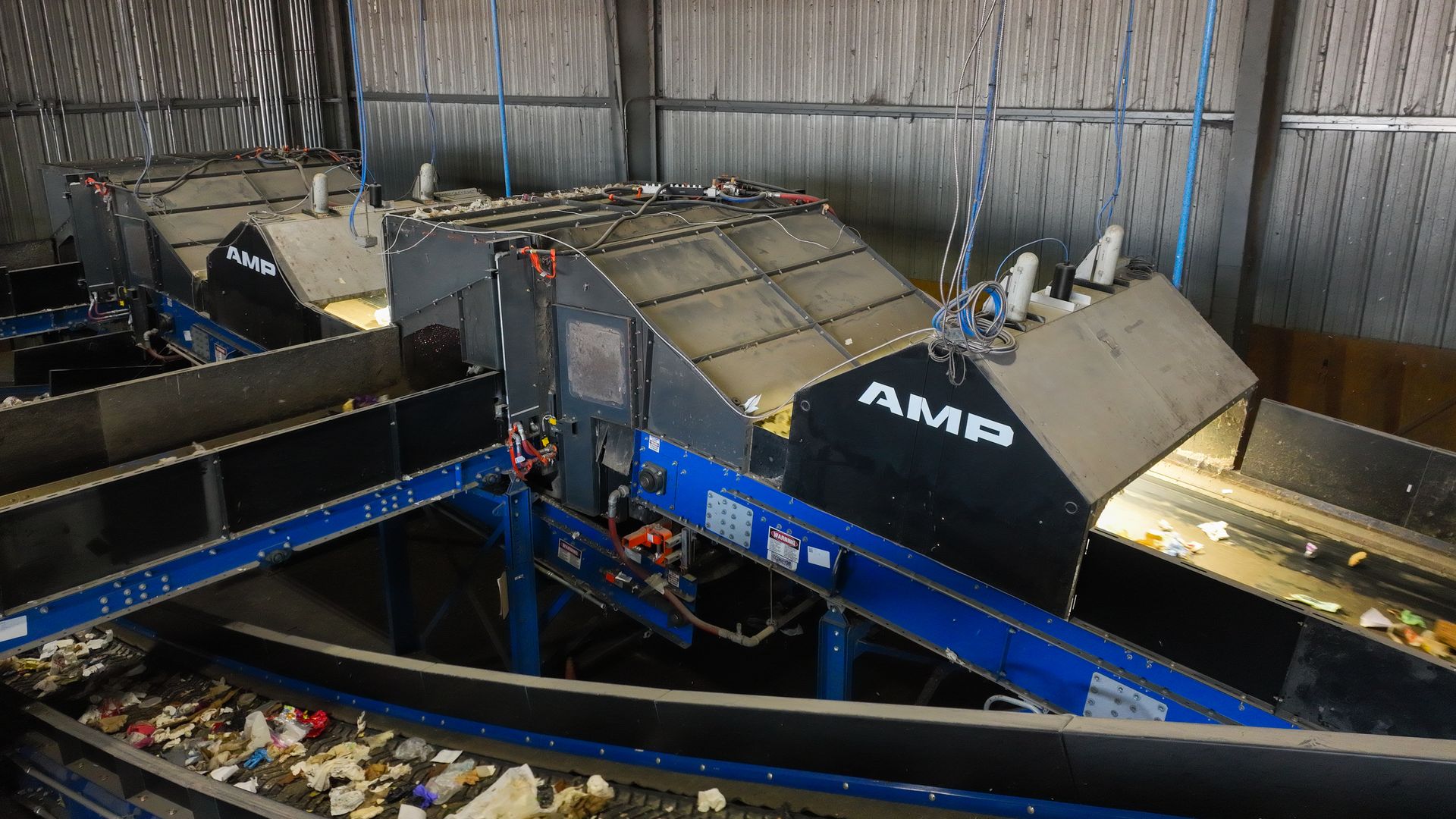 Industrial waste processing line inside a metal warehouse: angled gray conveyors on blue frames, tangled cables, and piles of trash on the belt; "AMP" logos on black panels.