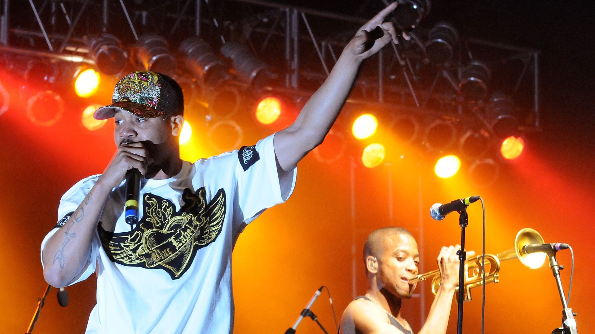 During an on-stage performance, Juvenile raps into a mic and Trombone Shorty can be seen behind him, playing his trombone into a microphone.