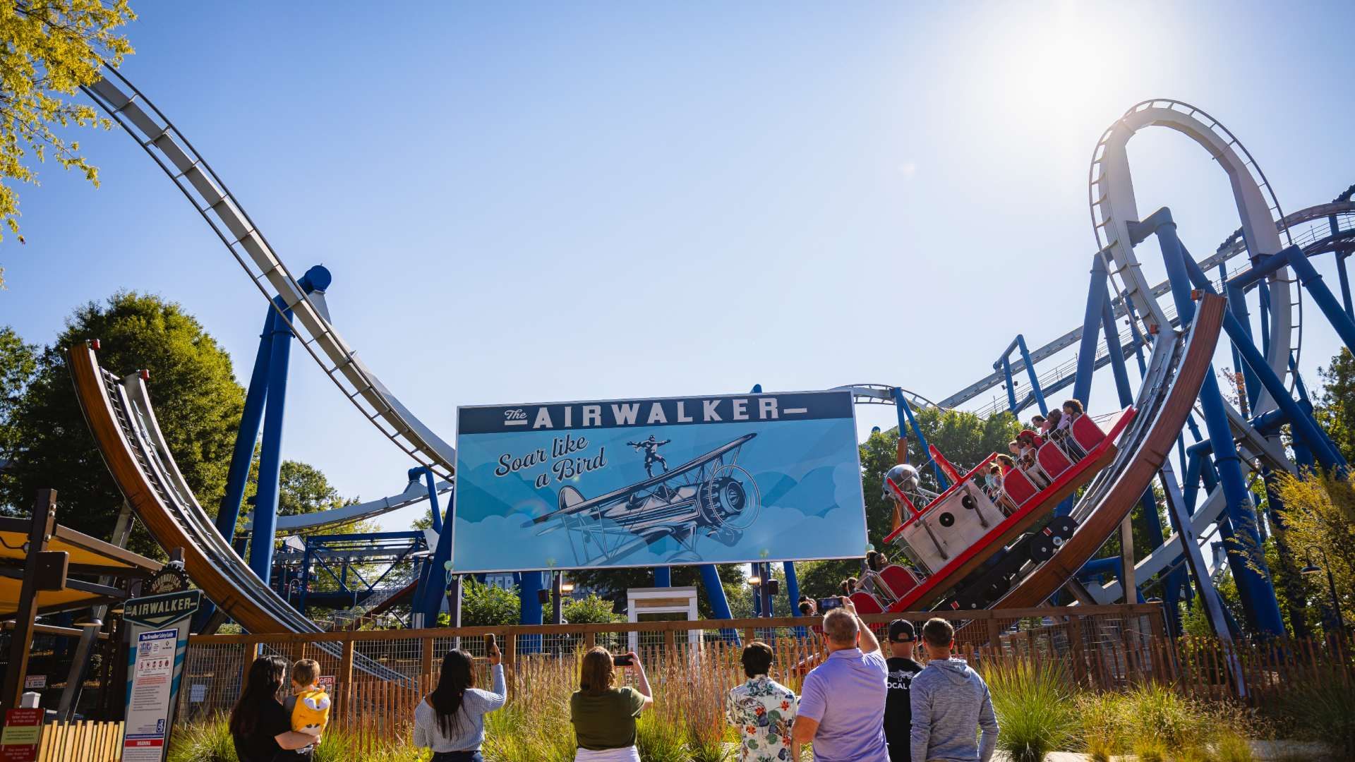Sunny day at an amusement park with a looping coaster called "The AIRWALKER." A blue sign reads "Soar like a Bird" and shows an airplane. People watch from the fence in the foreground.