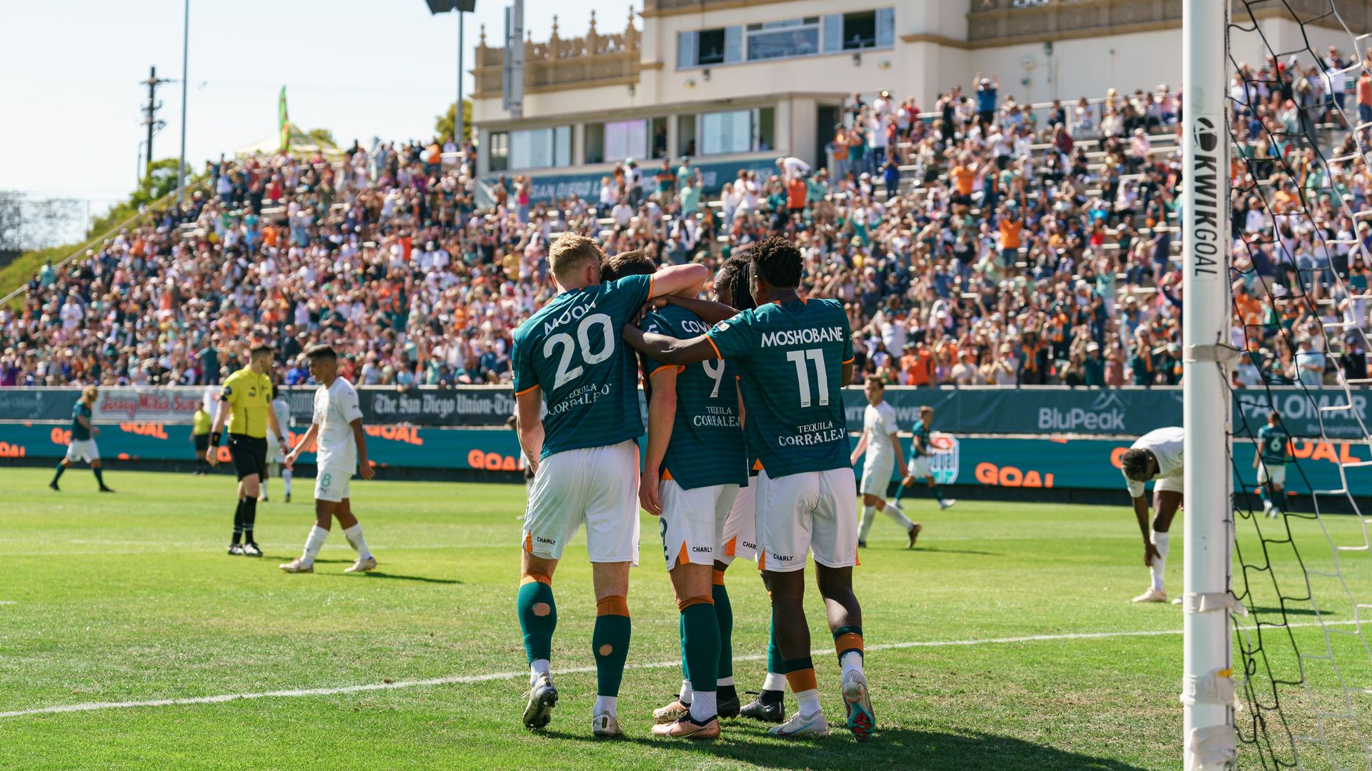 Men's soccer players in teal and orange jerseys hug each other on the field after a goal with fans in the stands. 