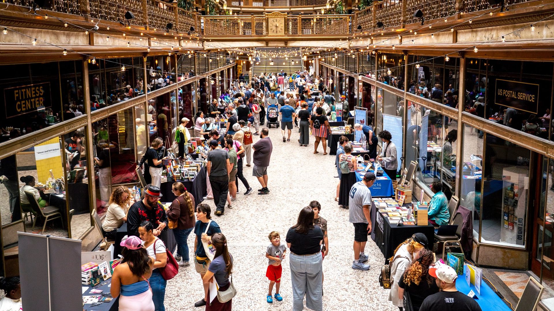 Bustling indoor book fair in a historic atrium with ornate gold railings and balconies. Strings of lights span the space as people browse tables full of books.
