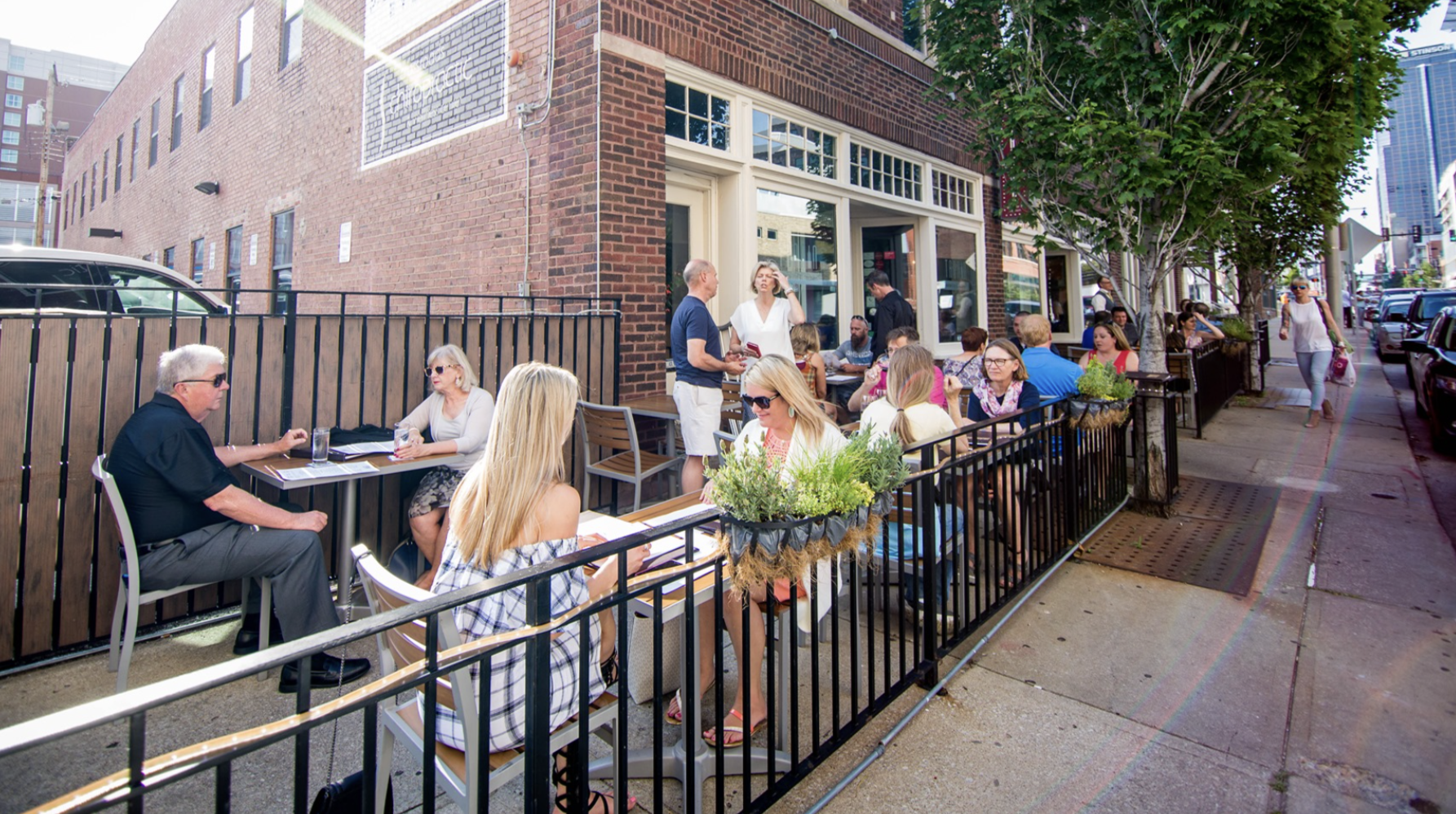 Outdoor sidewalk cafe along a brick building, diners at tables behind a black railing with hanging planters; sunlit street with trees and pedestrians in casual summer clothing.