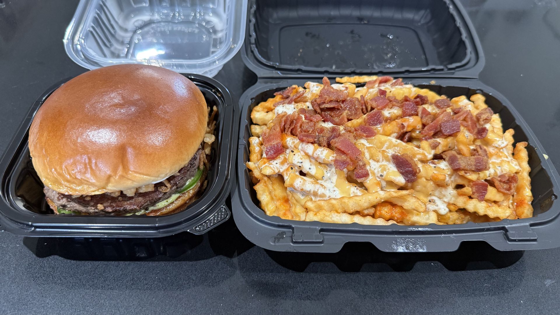 A burger in a black container next to crinkle-cut fries topped with cheese sauce and bacon bits in a black container.