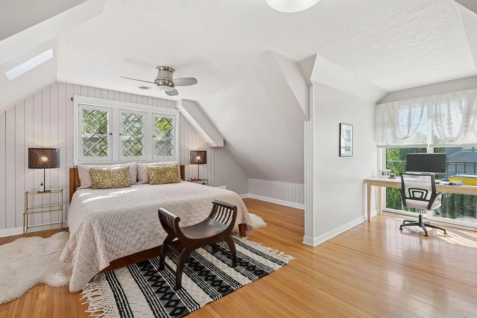 Bright bedroom with angled ceiling, striped bedspread, floral pillows, patterned rug, dark wood bench, lamps on gold stands, and a workspace by window with desk and chair.