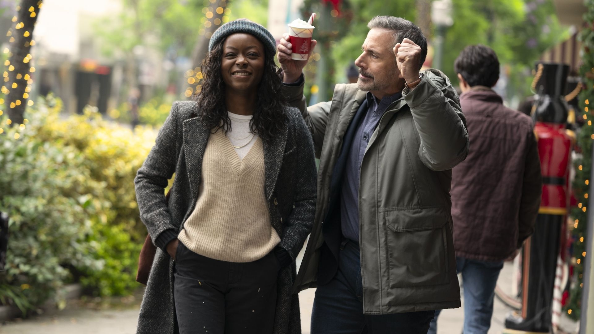 Smiling woman in gray coat and sweater walks on a decorated street with man in green jacket holding a cup with whipped cream and candy cane, raising fist in celebration.