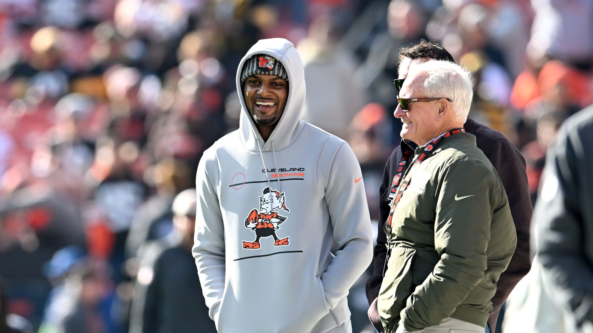 Two men smile at a crowded stadium: a man in a light gray Cleveland Browns hoodie and beanie, and another in an olive-green jacket wearing sunglasses, with blurred spectators in the background.