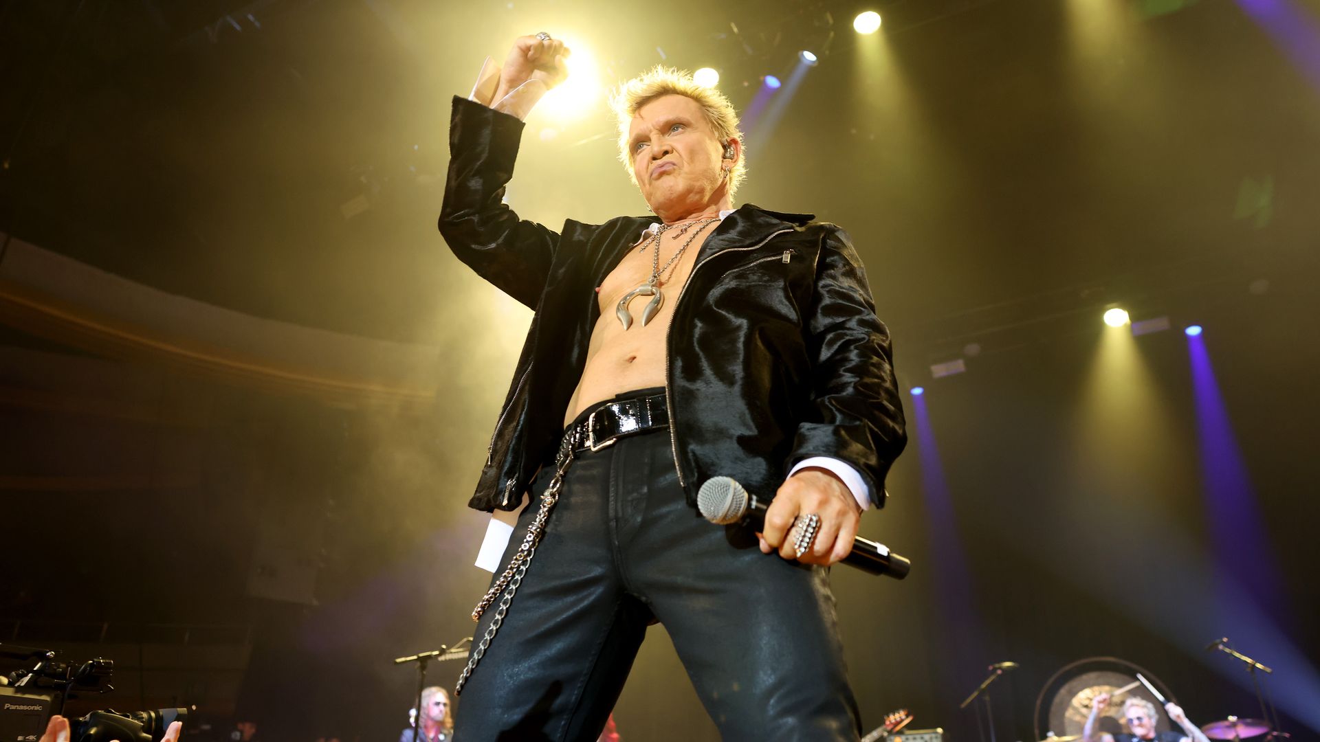 A male rock singer on stage raises his fist, wearing a shiny black jacket and necklace, microphone in his other hand. Yellow spotlights beam as a band plays in the background.