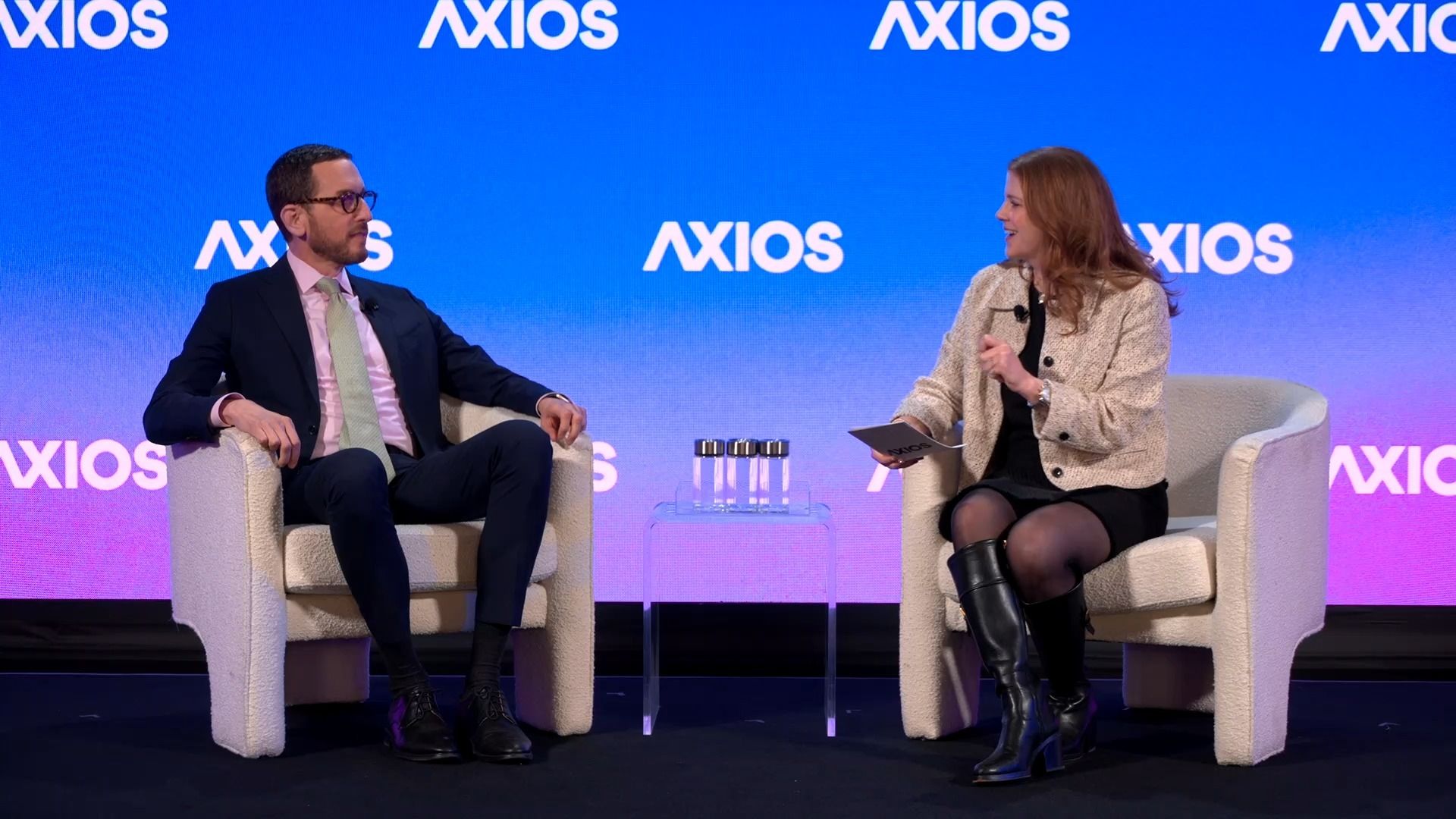 Two people sitting in white armchairs on a stage with blue and purple background displaying the word "AXIOS." The man wears a dark suit and light green tie, the woman a beige jacket and black boots.