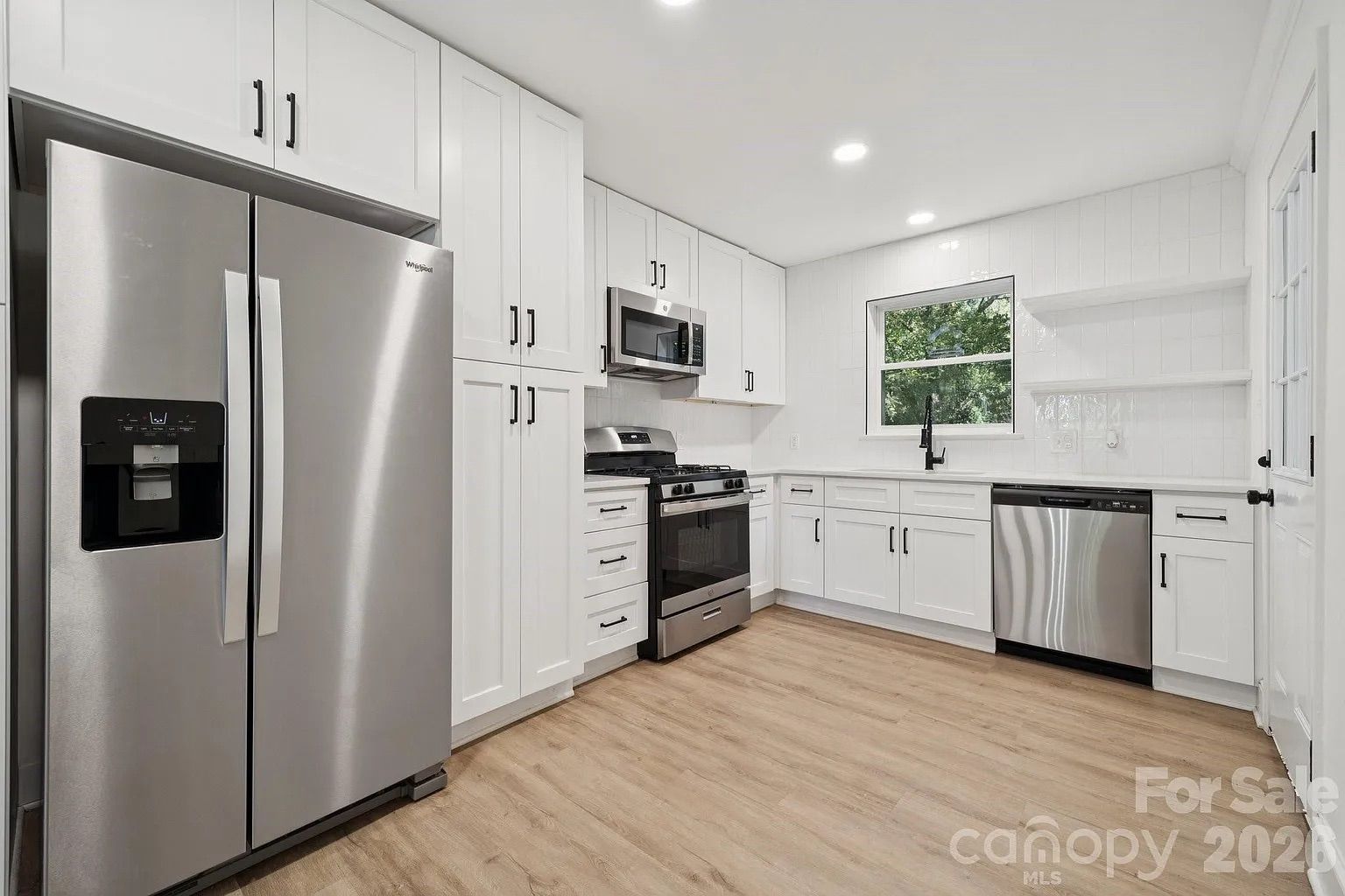 Modern kitchen with white cabinetry, stainless steel Whirlpool refrigerator, stove, microwave, and dishwasher, light wood flooring, black faucet, and a window showing green trees outside.