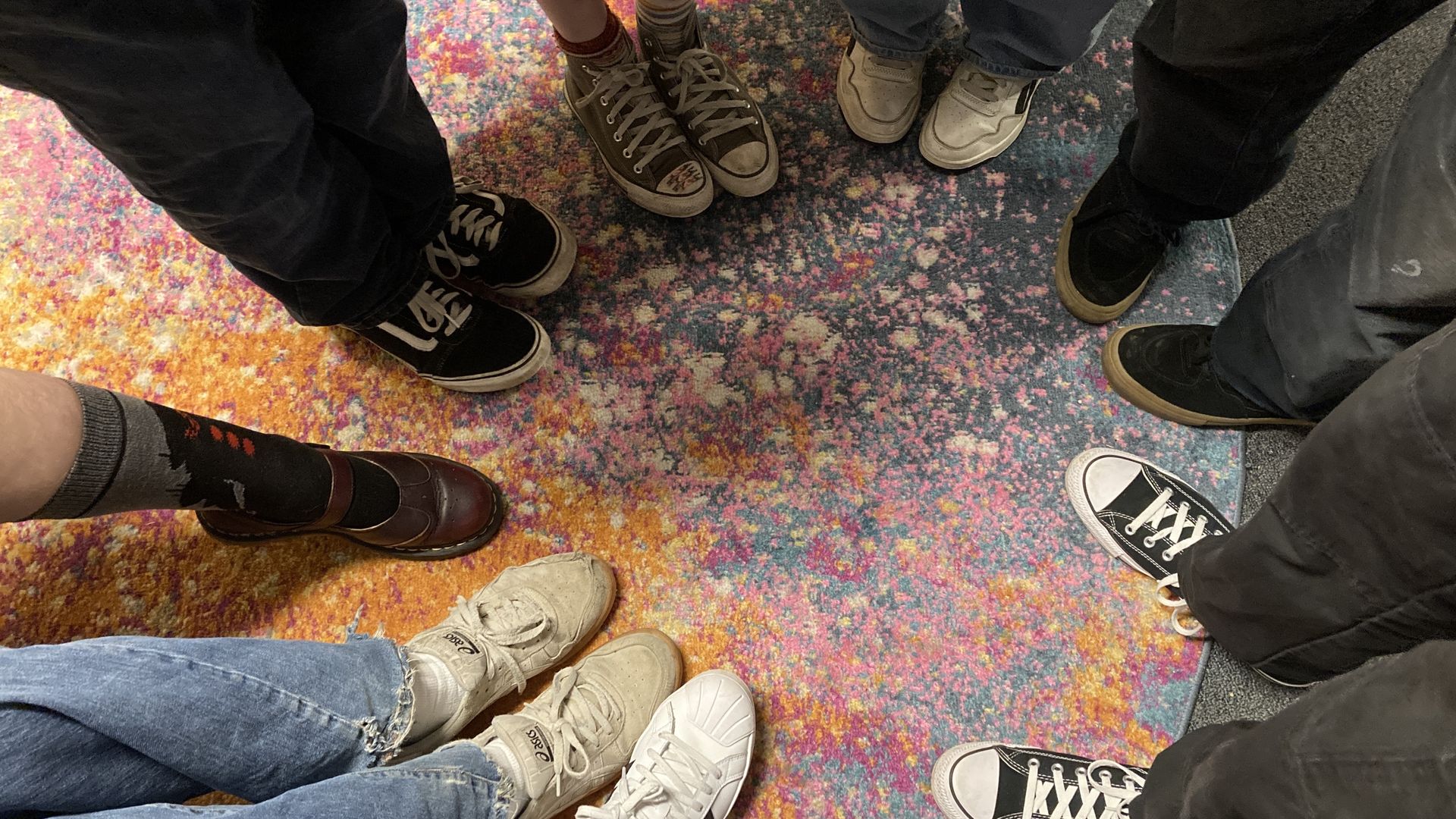 The feet of eight people, all wearing different shoes, in a circle on a multicolored carpet.