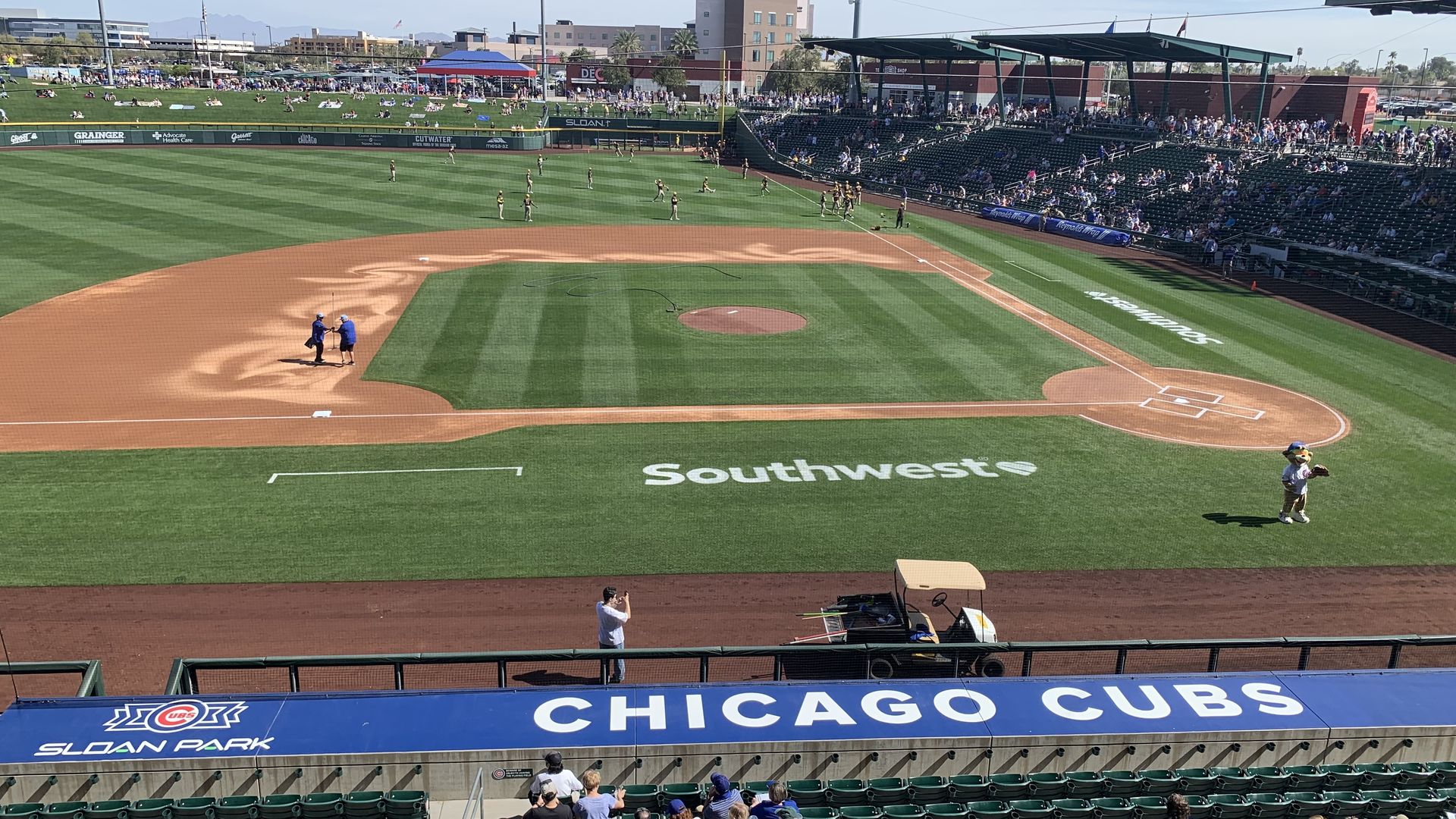 View of a sunny baseball field with green grass and brown infield at Sloan Park. Fans seated in stands behind outfield and along the first base line. Mascot near home plate area. "Southwest" logo visible on grass.