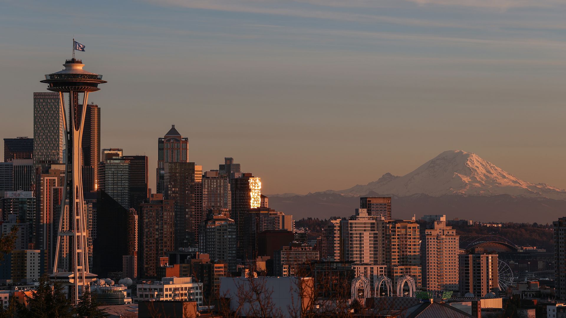 Sunset over the Seattle skyline with the Space Needle on the left, glassy high-rises bathed in orange light, and a distant snow-capped mountain on the horizon.