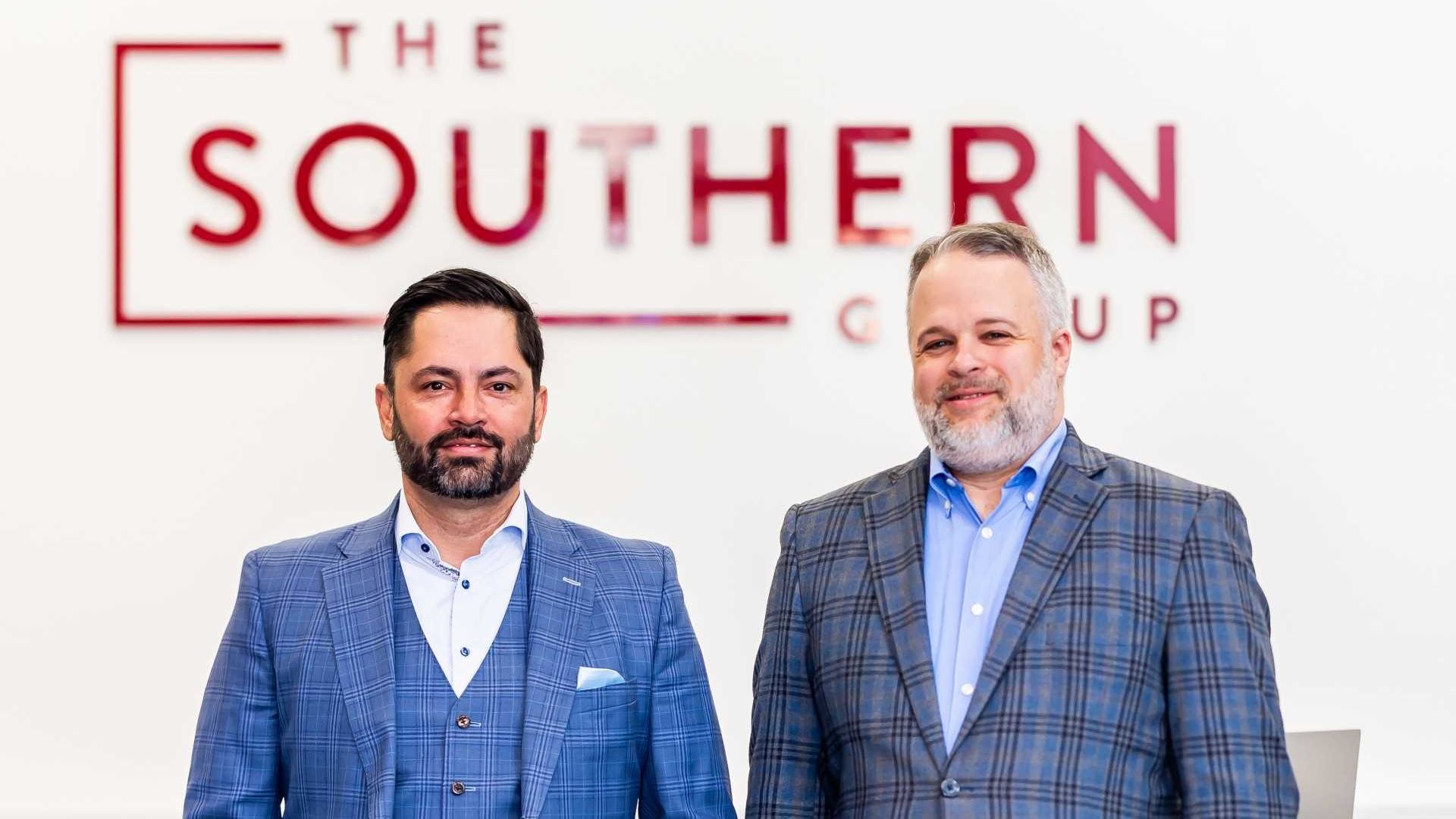 Two men in blue plaid suits standing with hands in pockets in front of a white wall and desk with a red sign that reads "The Southern Group".
