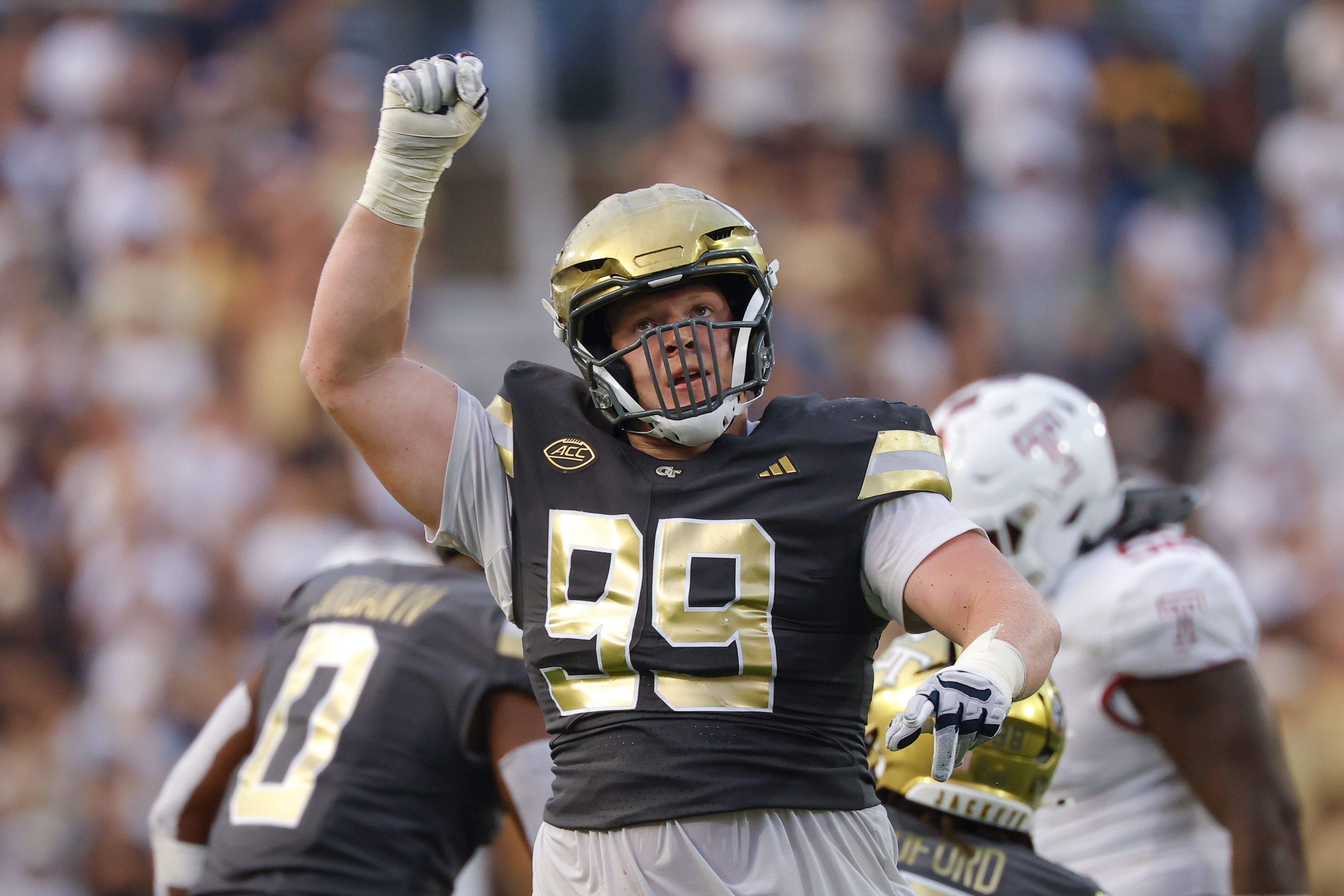 A football player in a black and gold uniform and gold helmet raises a gloved fist in celebration on the field, with a blurred crowd in the stands behind him.