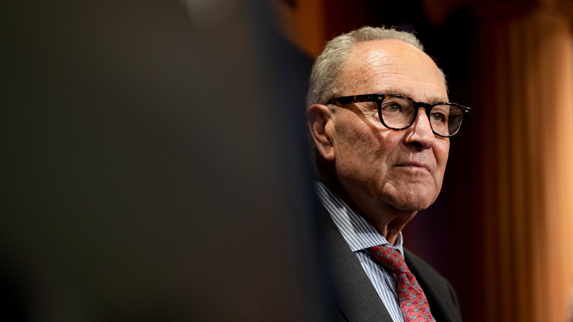 Chuck Schumer wears glasses, a red tie, a striped blue and white shirt and a dark suit as he looks on during a press conference.