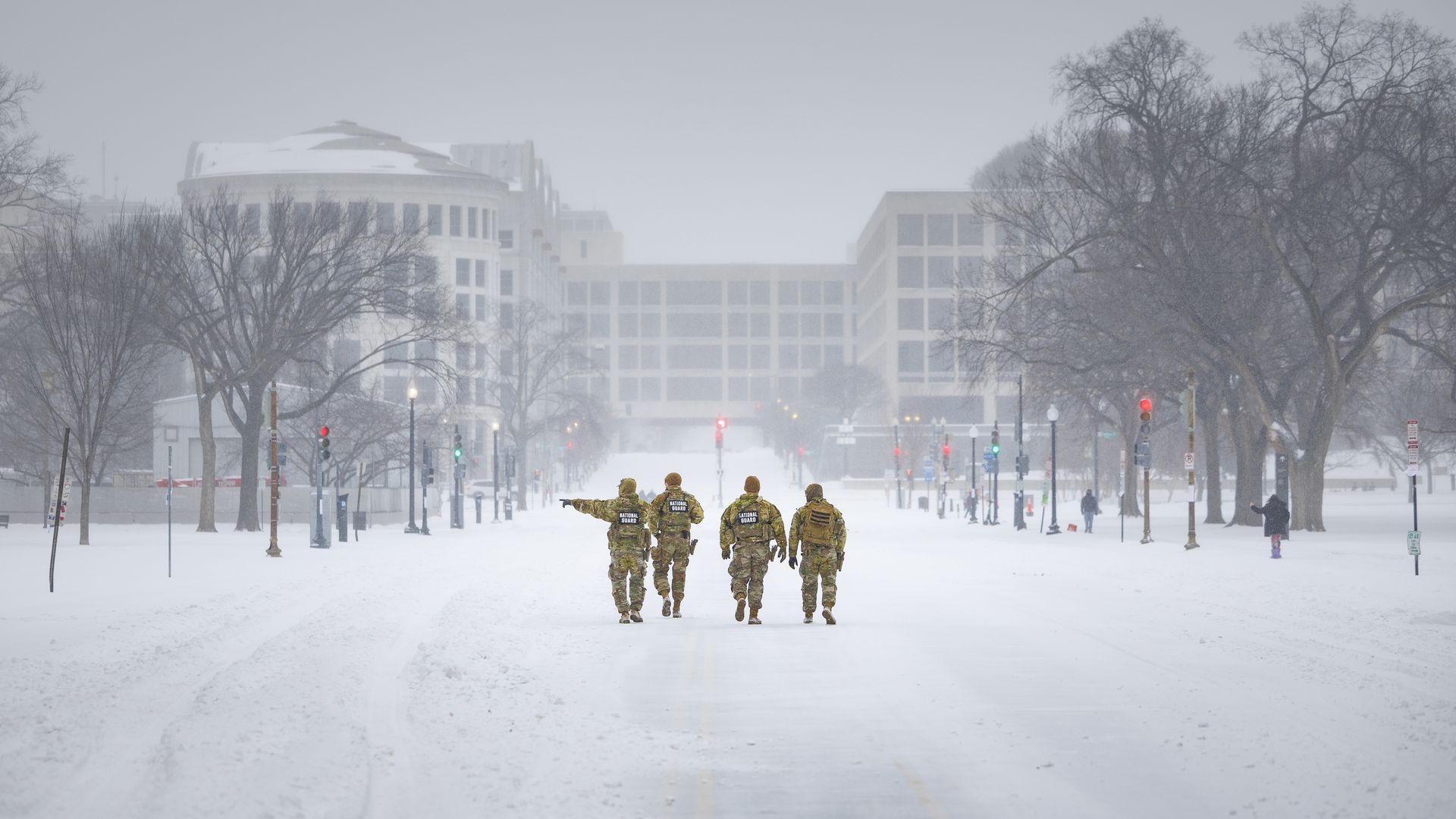 Four National Guard soldiers in camouflage uniforms walking on a snow-covered street in a city with leafless trees and buildings in the foggy background.