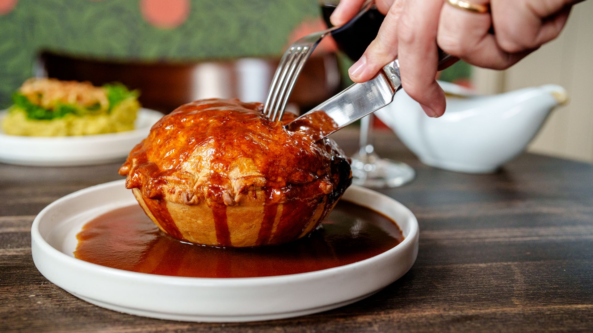 Someone uses a knife and fork to cut into a braised short rib pie, which has been cooked to golden brown, and is covered with a dark red sauce.