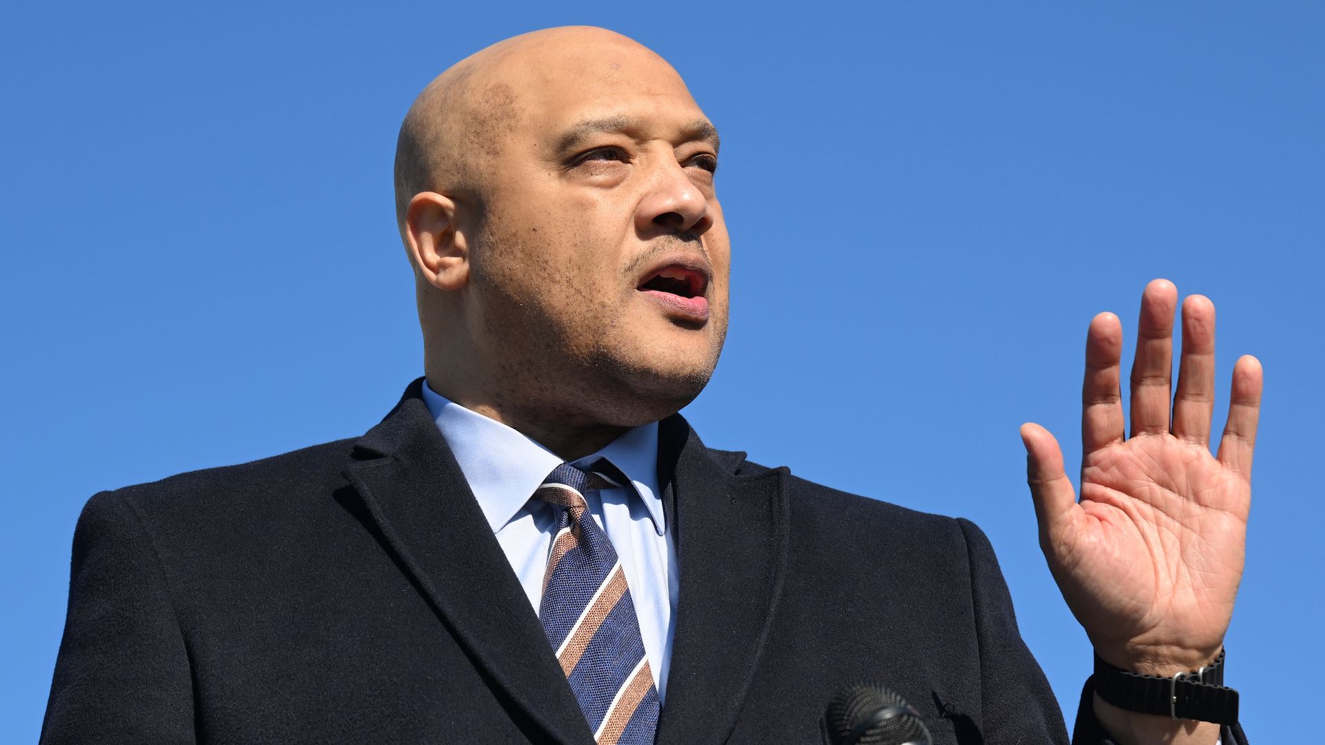 A bald man wearing a suit and tie speaks into multiple microphones outdoors against a clear blue sky.