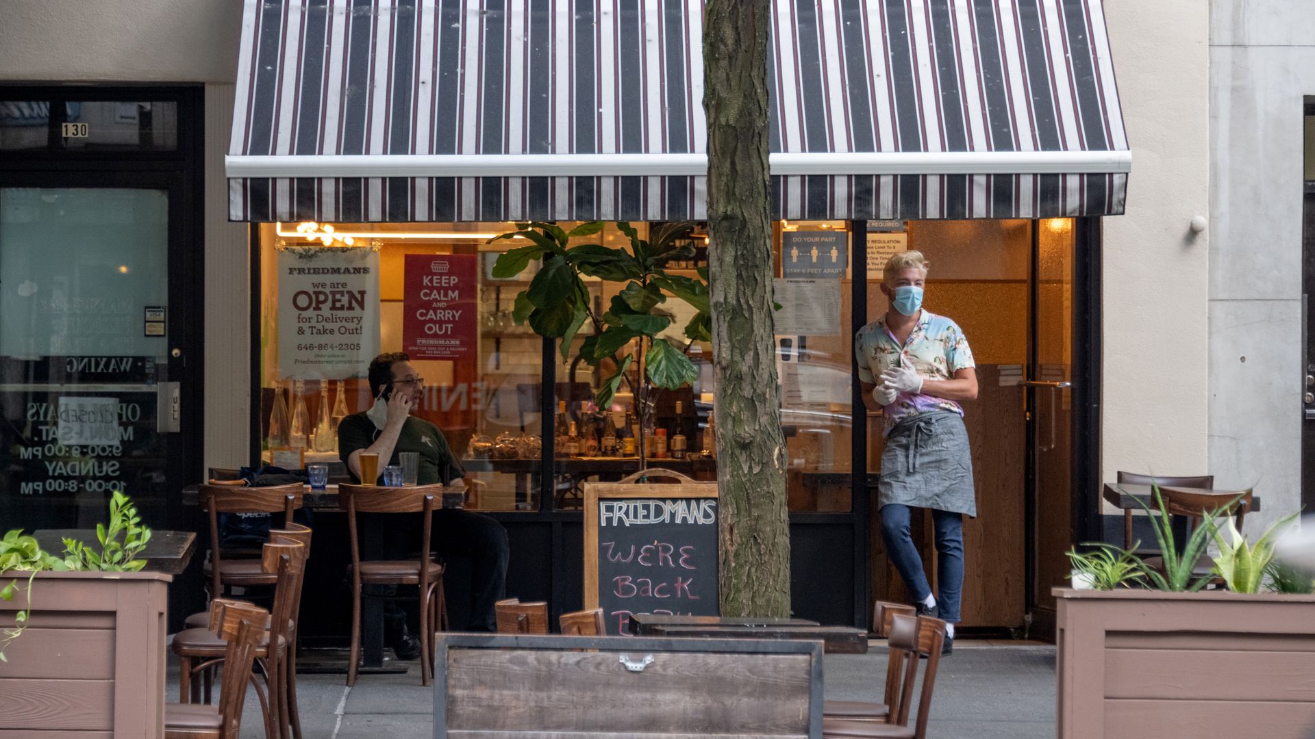 A waiter stands in the doorway of a restaurant in New York City while wearing gloves and a face mask