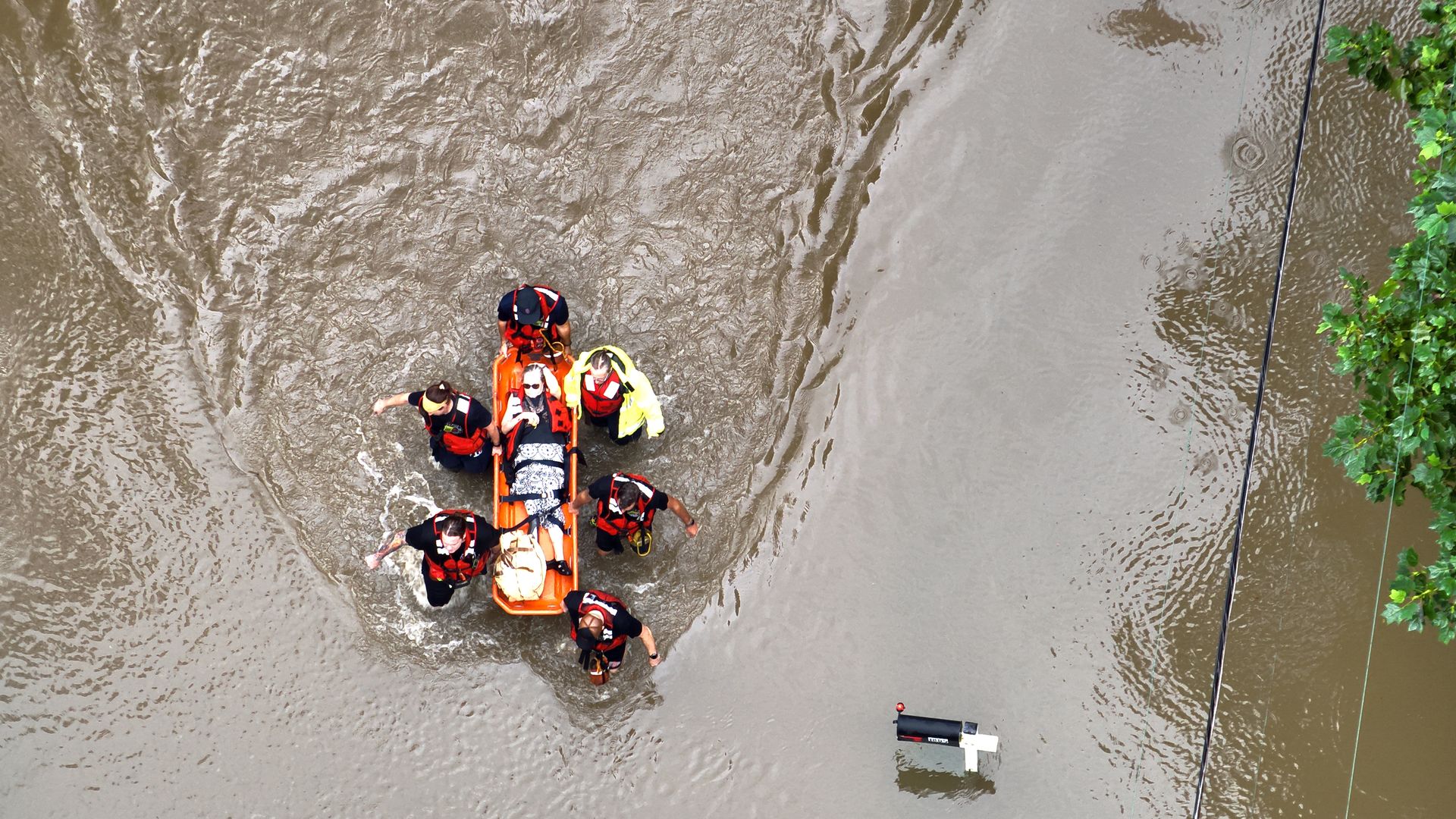 Aerial view of a rescue team in orange life jackets on an orange inflatable boat in muddy floodwater, with a bright yellow STOP AHEAD sign on a post and green foliage along the right edge.