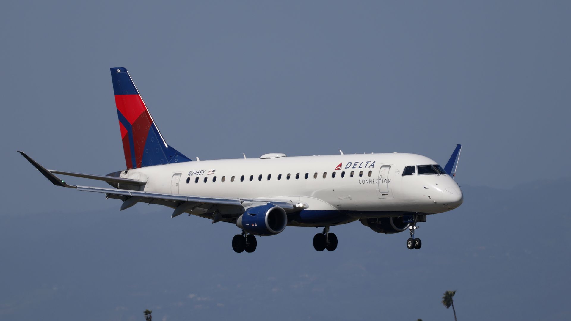 A Delta regional jet with white fuselage, blue tail with red accent, descending with landing gear down. Underwing engine visible; blue sky and desert foreground with a few palm trees.