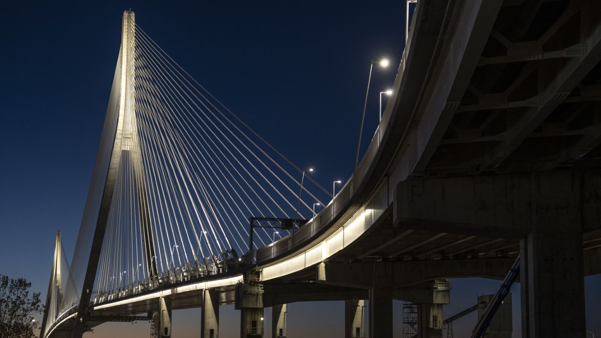 Illuminated cable-stayed bridge at dusk with a dark blue sky and streetlights along the curved roadway. Industrial structures visible beneath and in the background.