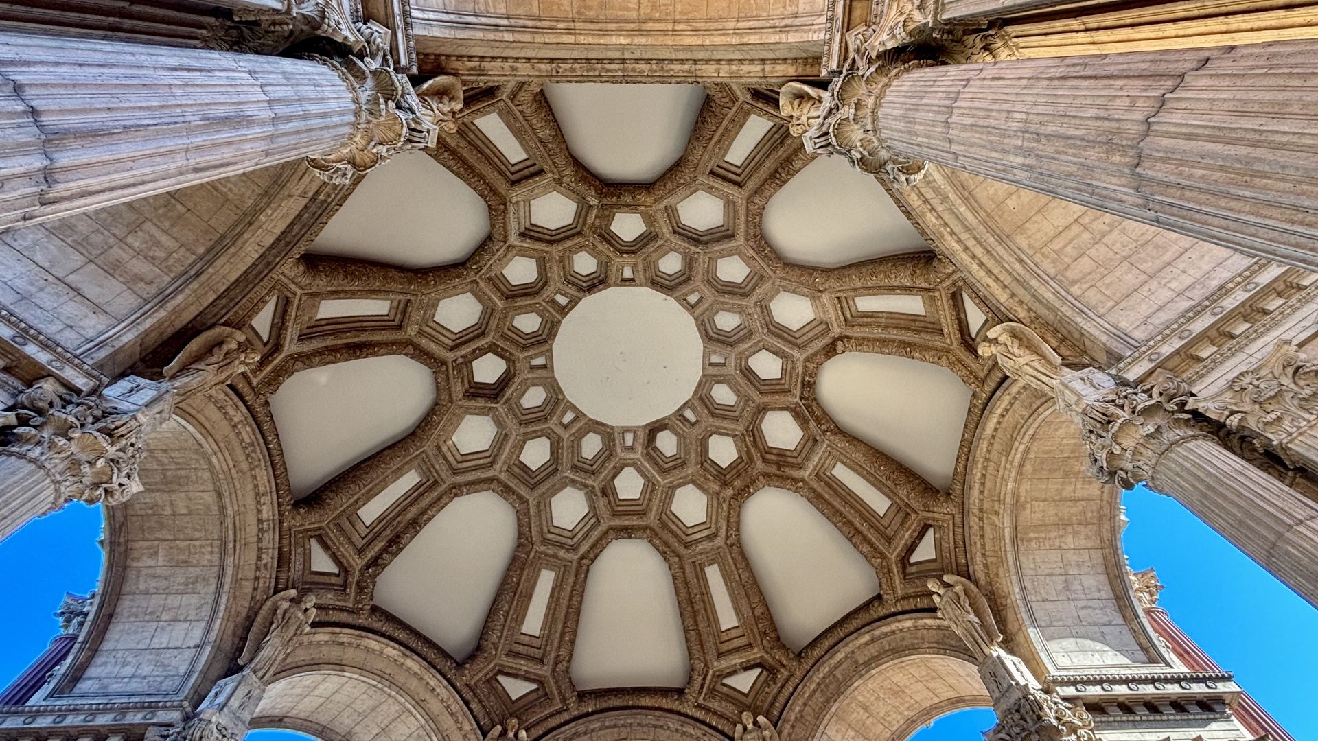 Looking up at a grand stone ceiling with hexagonal coffers, ornate arches and columns, and sculpted figures; blue sky shows through the openings.