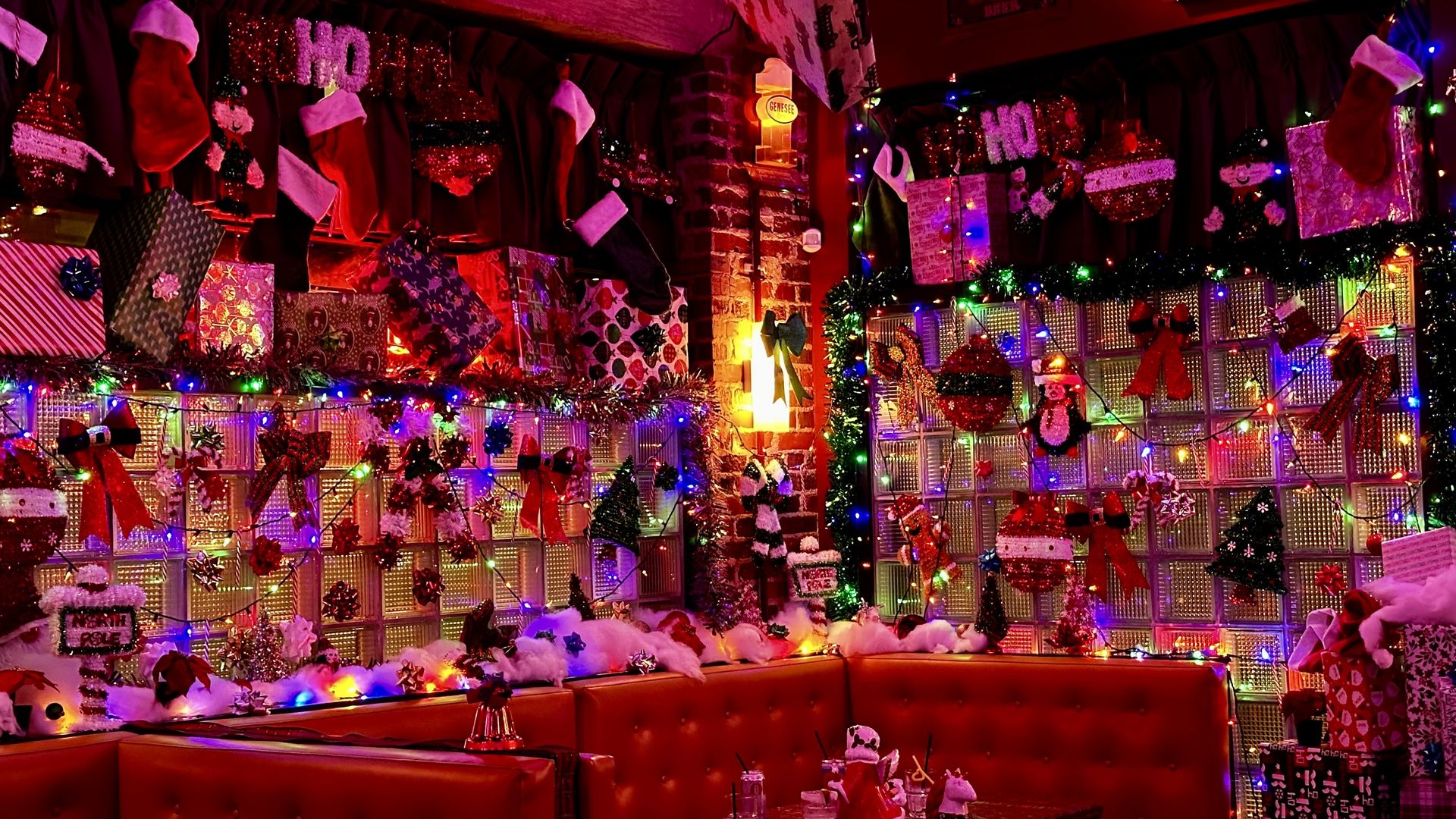Cozy booth area decorated with red curtains, stockings, wrapped gifts, colorful Christmas lights, bows, ornaments, and holiday figures on glass block walls.