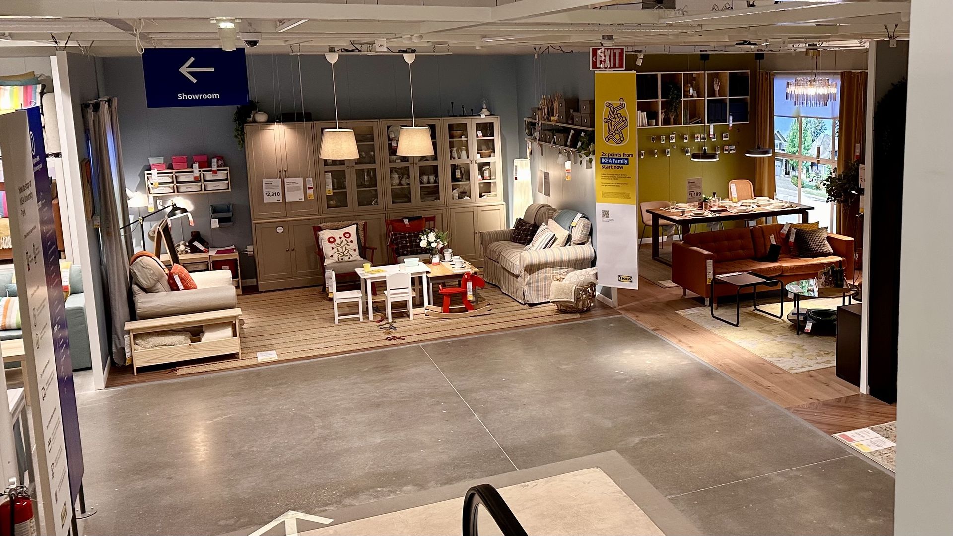 Indoor furniture showroom scene with gray-blue walls, glass display cabinets, beige sofa and plaid chair, a small white dining set, hanging lamps, and a yellow accent wall by large windows.