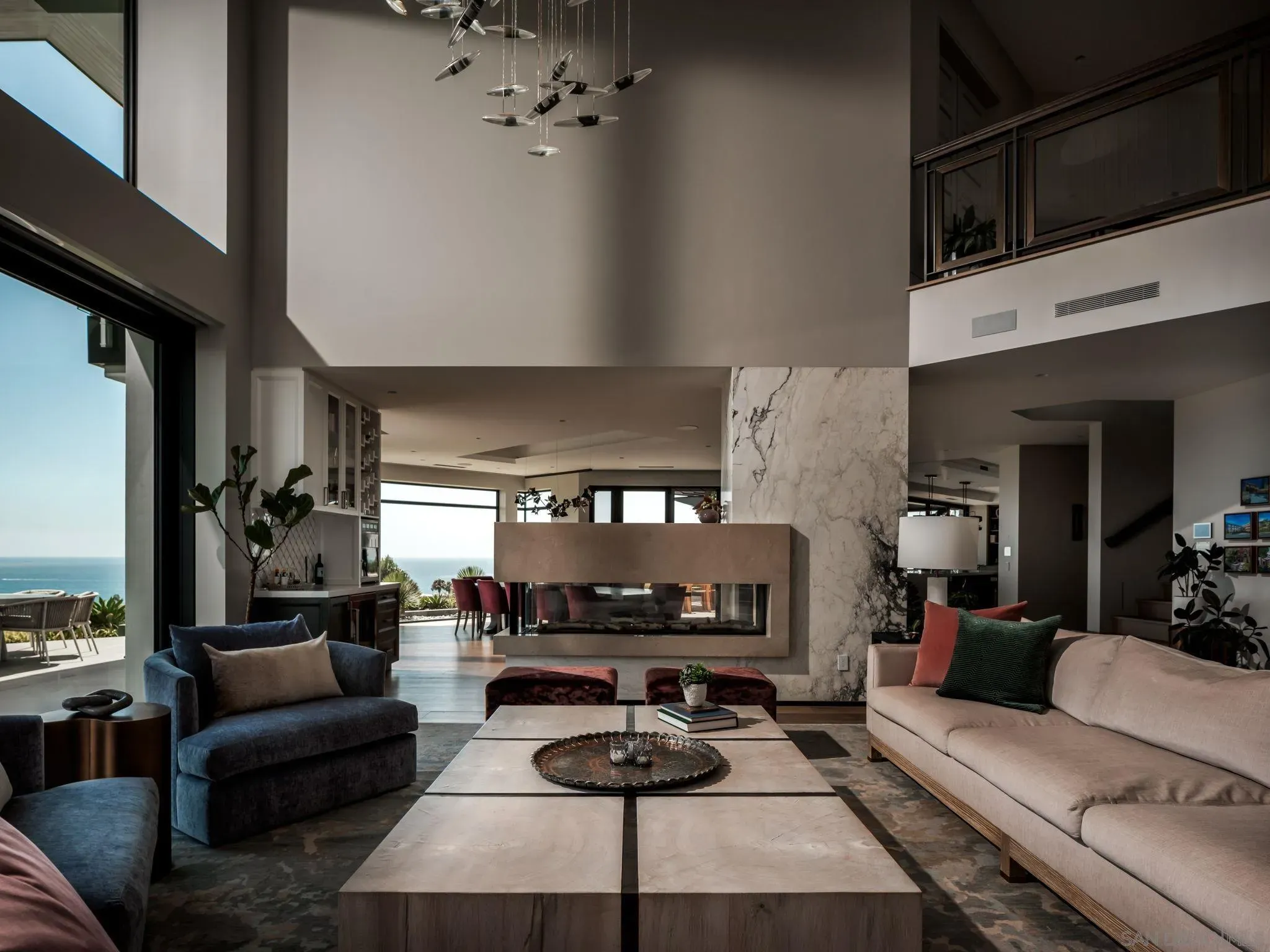 Spacious modern living room with marble surfaces and floor-to-ceiling windows overlooking the ocean. A sculptural chandelier of suspended discs hangs above beige and blue seating, with plants nearby.