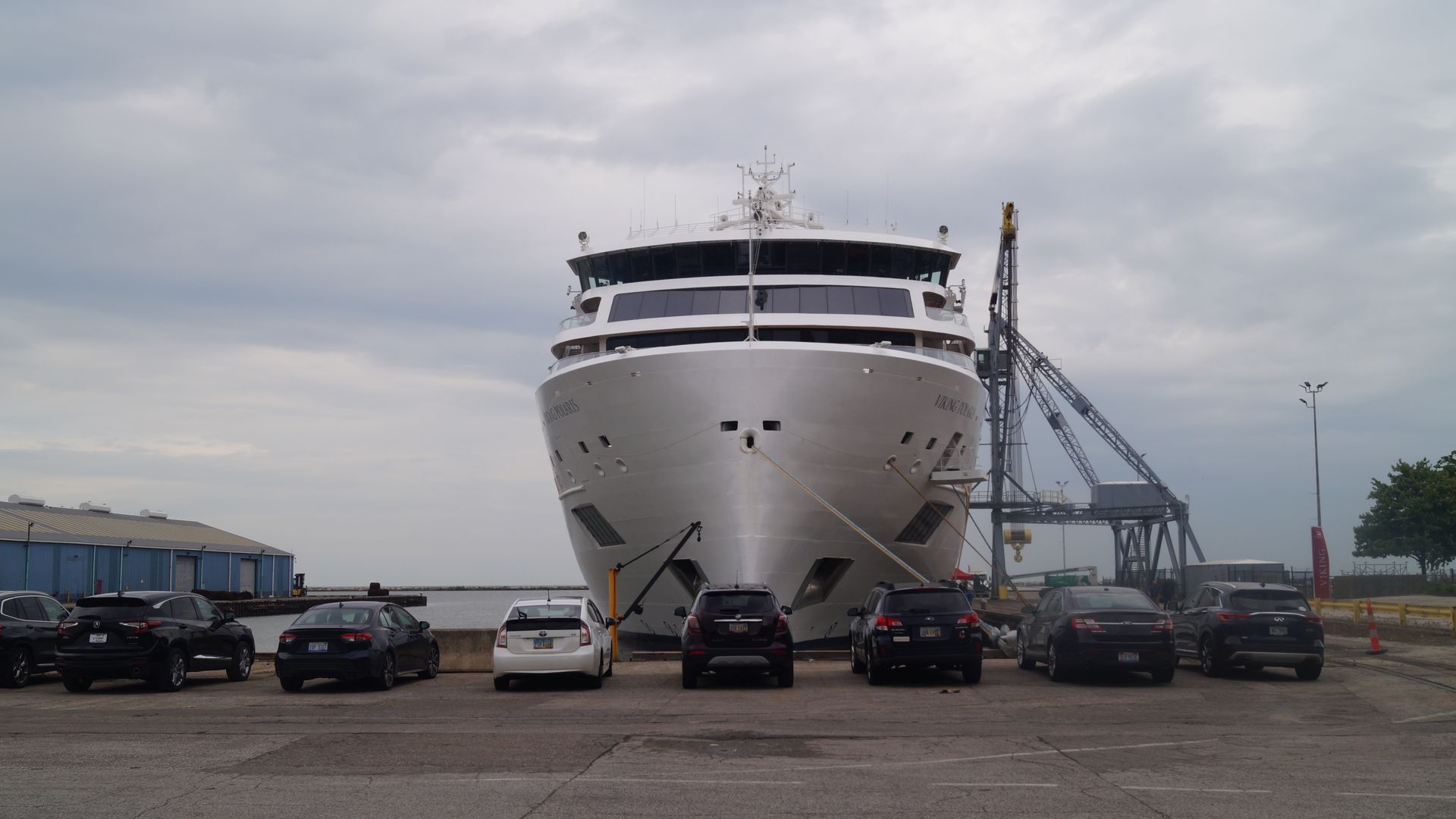 Front view of a large white cruise ship docked at a pier with several parked cars in front on a cloudy day, with industrial buildings and a crane nearby.