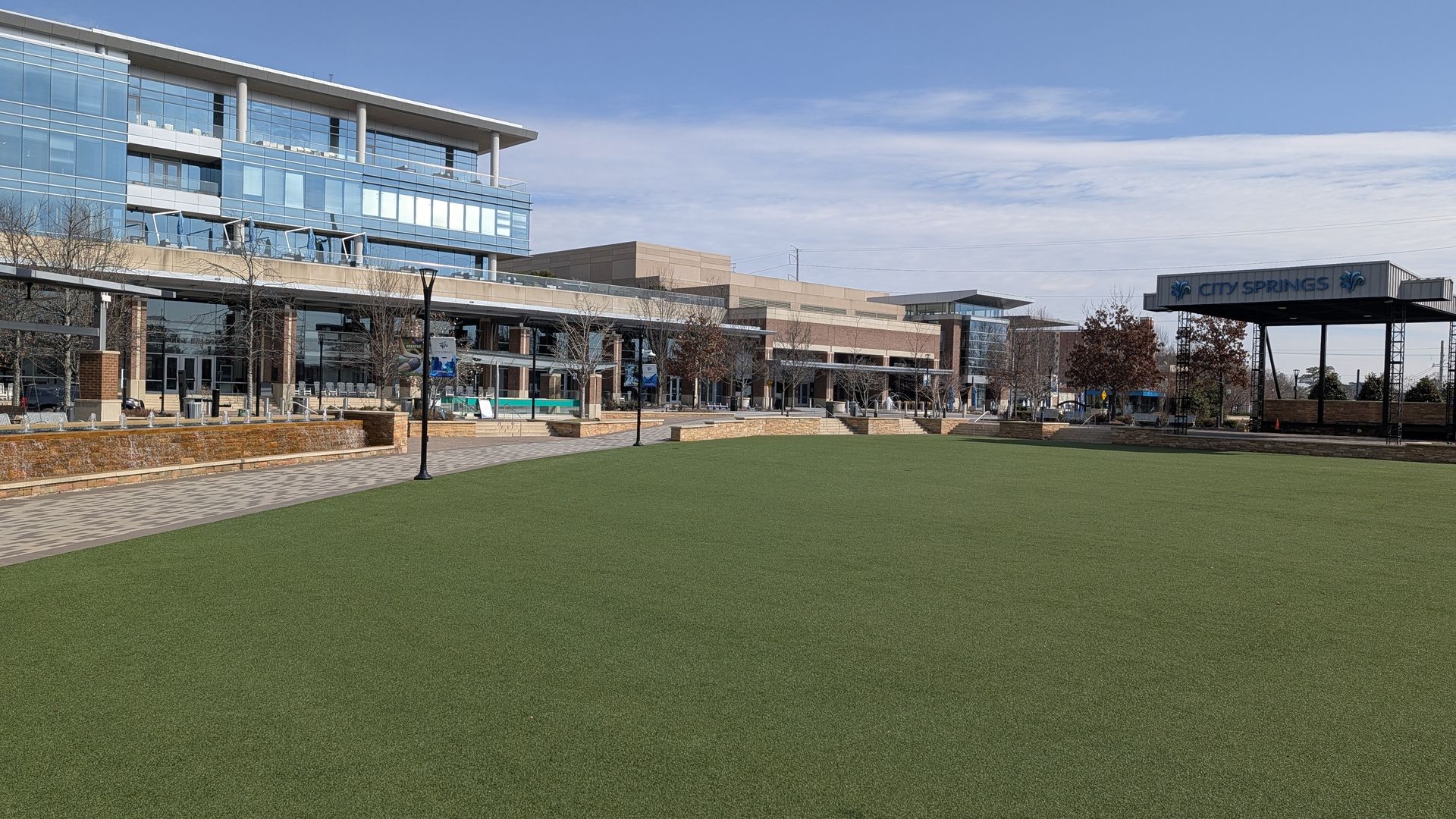 Open green park area in front of modern glass and brick buildings under a blue sky with a sign reading City Springs on a stage structure to the right.