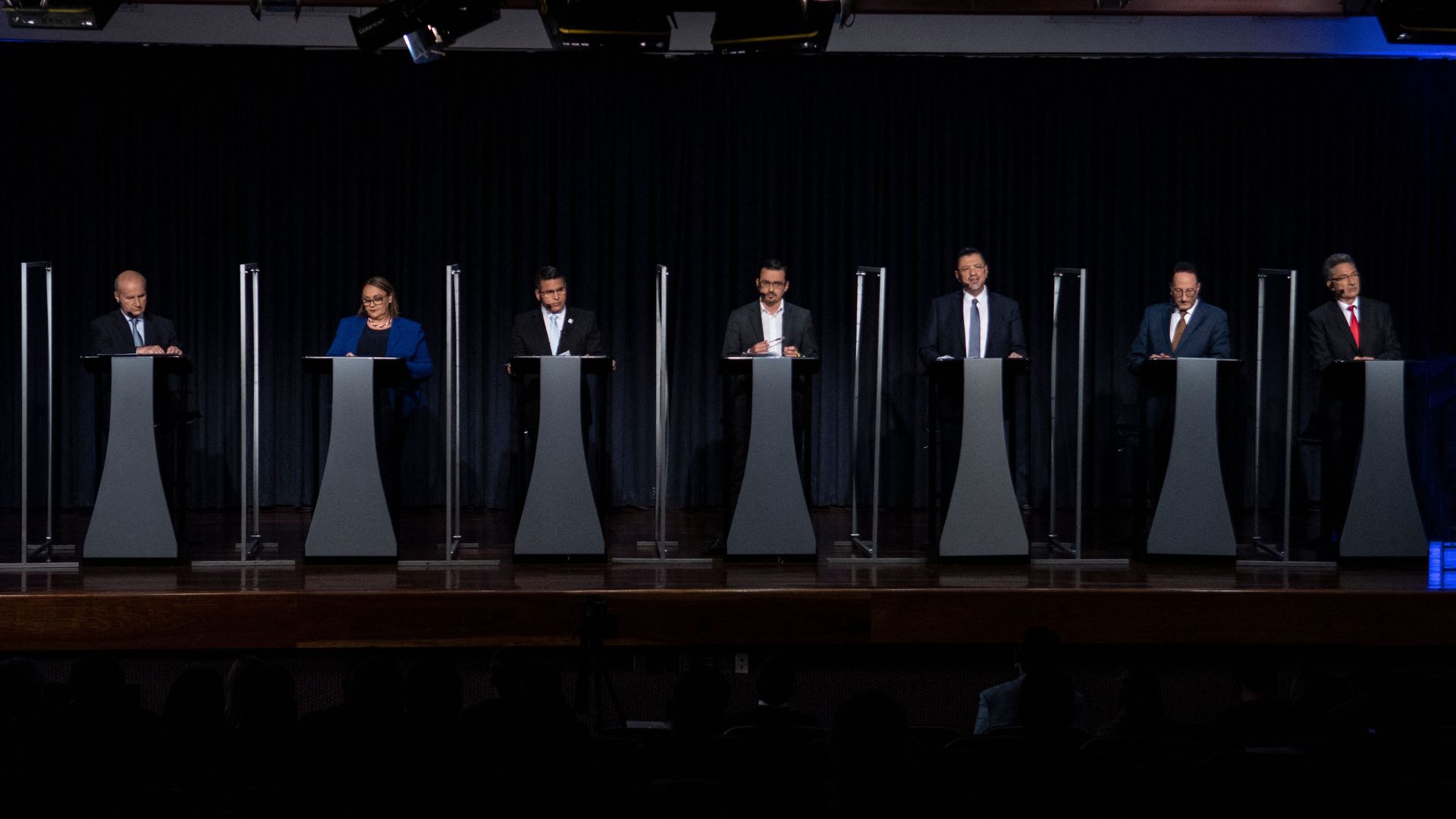 Six men and one woman who are all running for president of Costa Rica stand in front of podiums during a debate in a darkened room with a black background