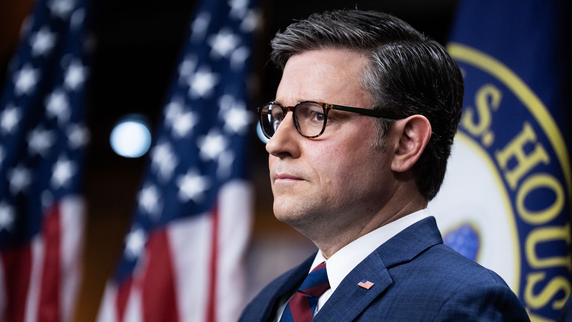 House Speaker Mike Johnson, wearing a blue suit, white shirt and blue and red striped tie, standing in front of flags.