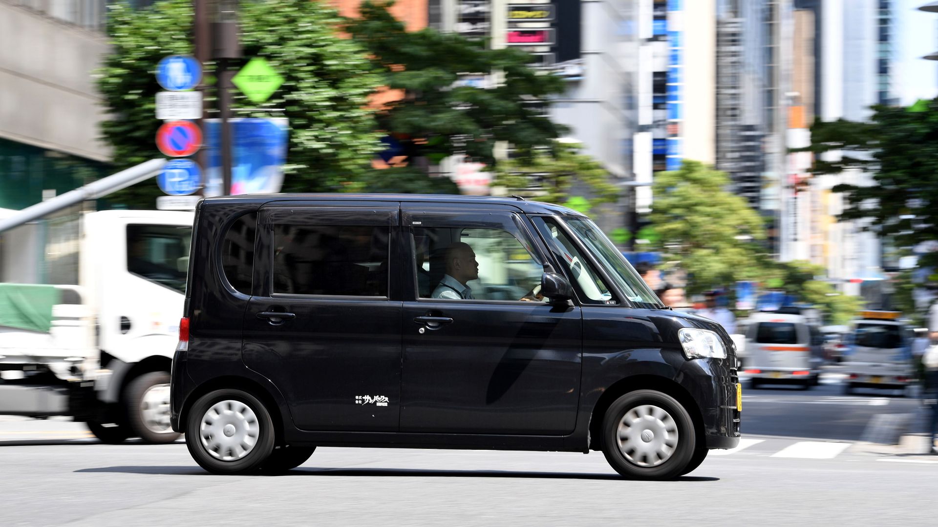 A black tiny kei car driving on a busy street in Japan, with the background blurred. 