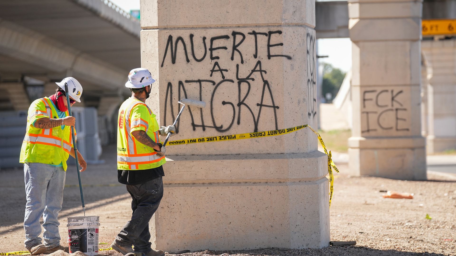 Two construction workers in neon safety vests and white hard hats paint graffiti on a large concrete pillar at a construction site, with yellow caution tape nearby and more graffiti elsewhere.