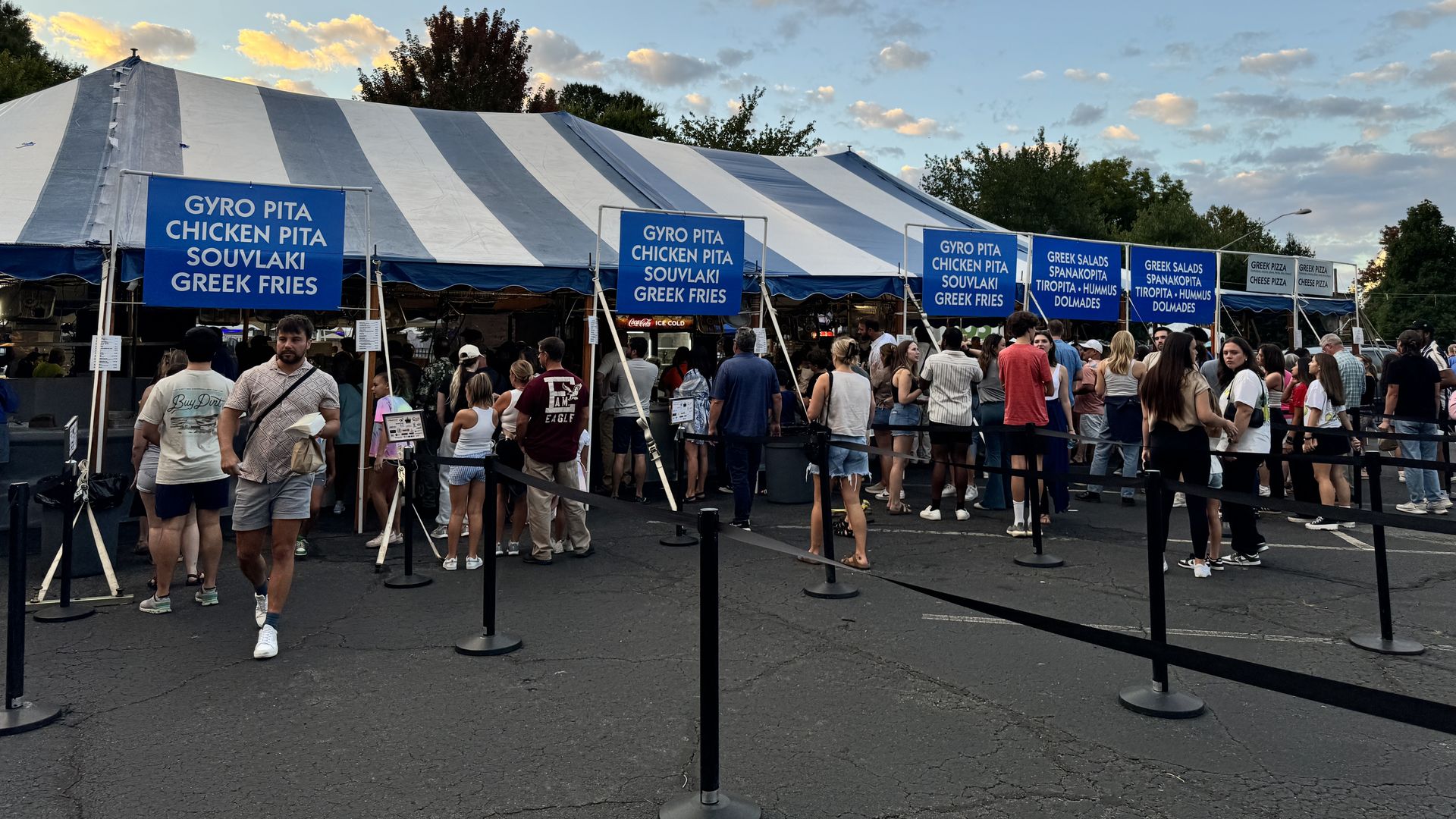 Greek Festival. Busy outdoor festival scene with a large blue-and-white striped tent. Blue banners list foods like gyro pita, chicken pita, souvlaki, Greek fries as people wait in line under a cloudy evening sky.