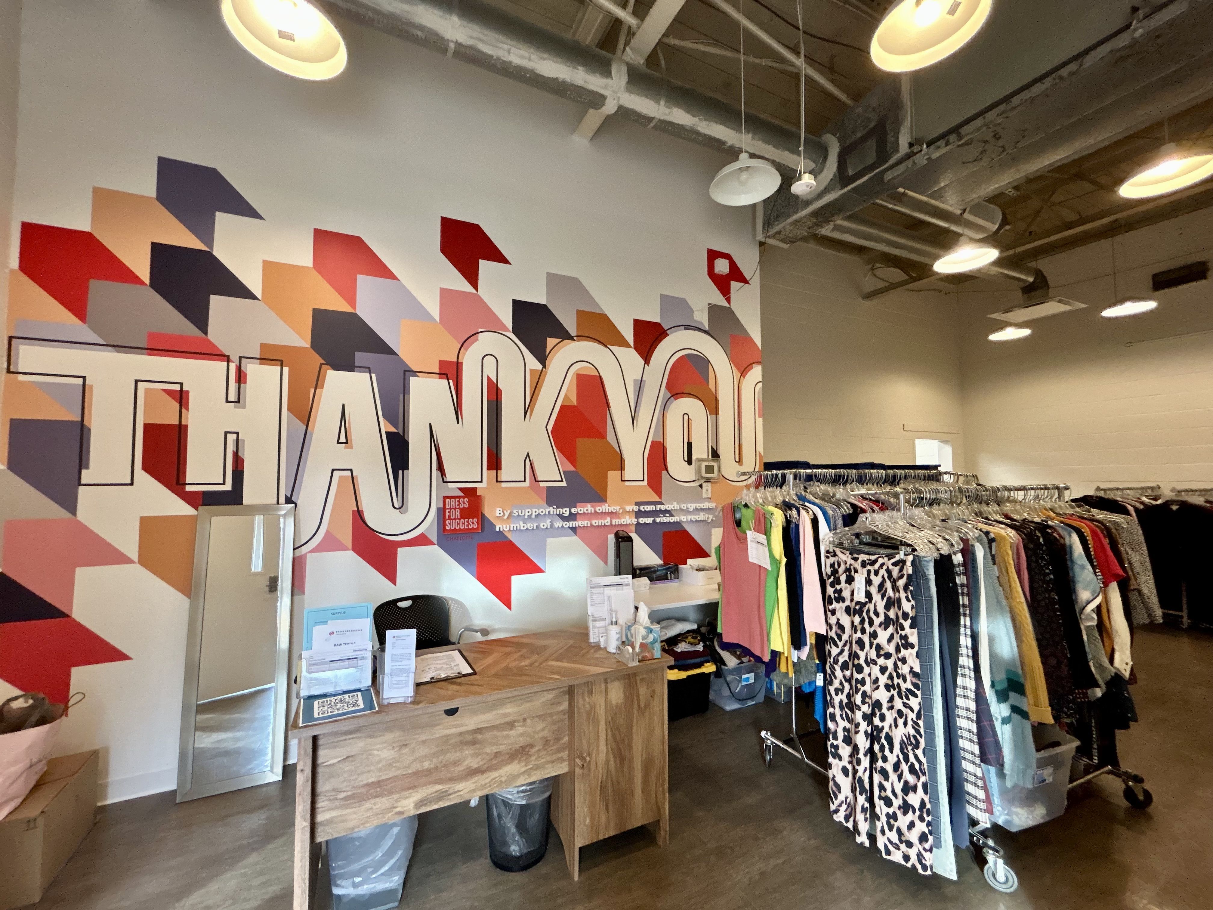 Interior of a clothing store with a wooden desk and mirror, colorful geometric wall art saying THANK YOU, and racks of assorted clothes including patterned pants and jackets under industrial ceiling lights.
