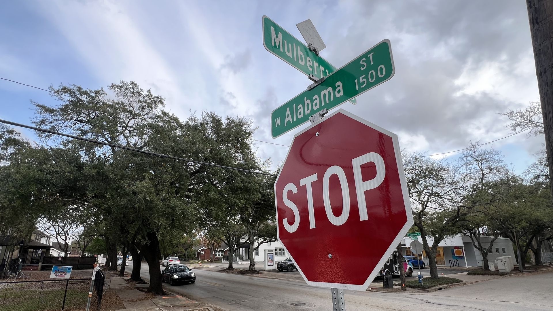 A street corner with a large red stop sign in the foreground; green street signs read Mulberry St and W Alabama 1500. A residential area with trees, parked cars, and a cloudy sky.