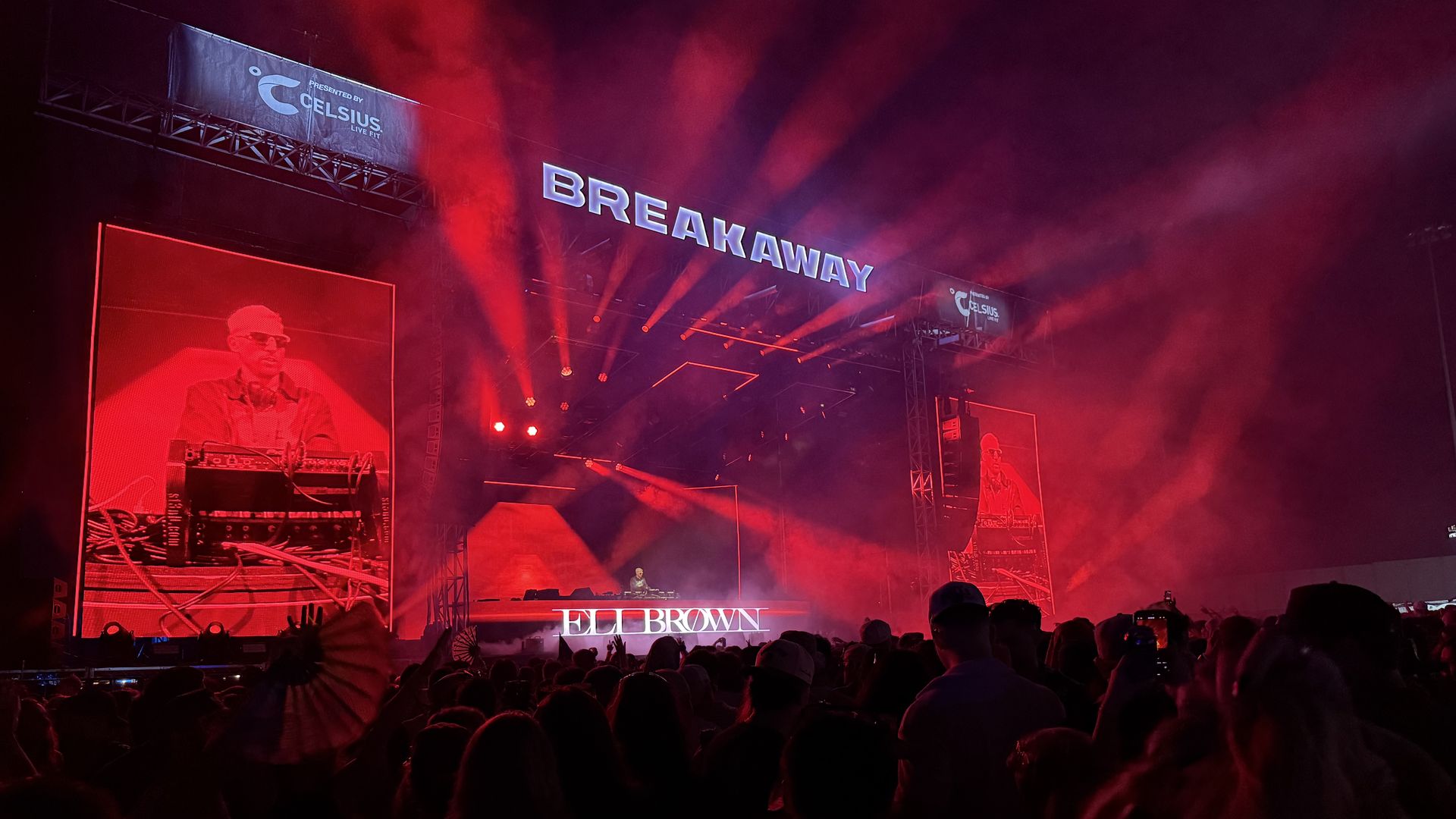 Wide concert scene with a large "BREAKAWAY" sign above a foggy stage. Red lights wash the crowd as screens show a DJ at a mixing desk; fans raise their hands.