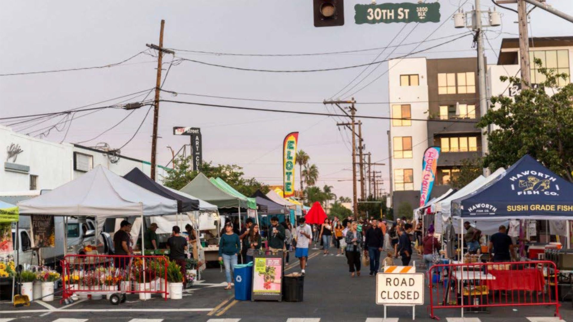 Street market on 30th St with vendor tents selling flowers, elote, sushi-grade fish, and ice cream. People walk between stalls on closed road under a cloudy sky near tall buildings.