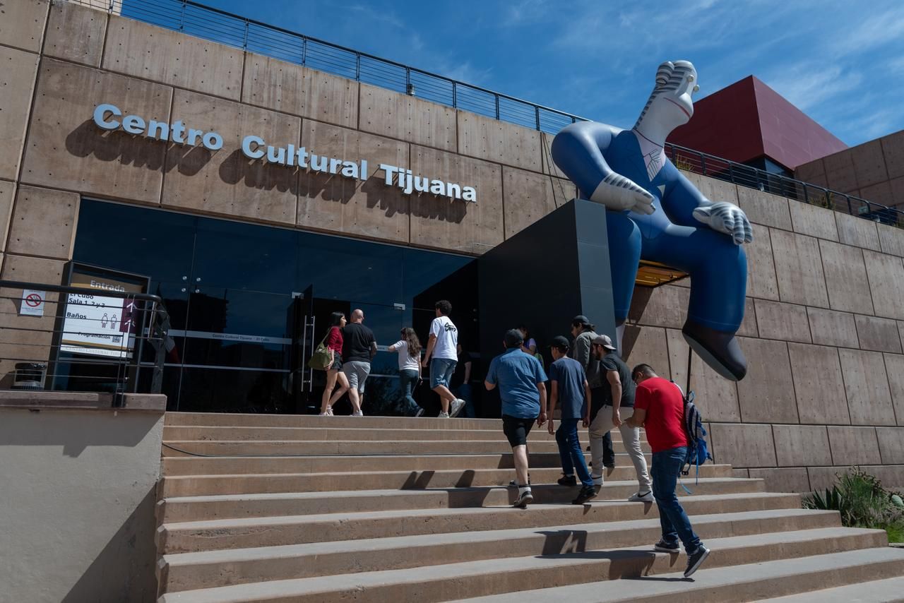Exterior of Centro Cultural Tijuana with a large blue statue seated at the entrance. People walk up beige concrete steps toward glass doors under a clear blue sky.